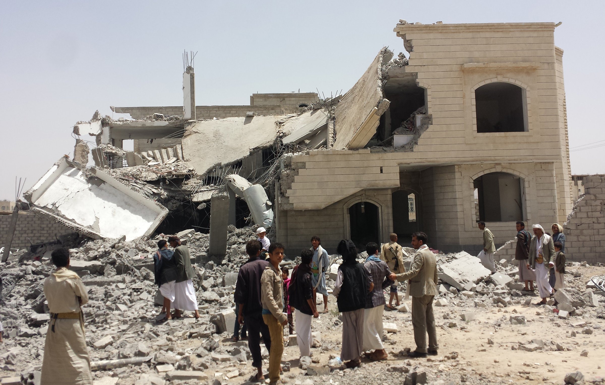 L'image montre un bâtiment partiellement détruit, avec des débris éparpillés autour. La structure semble avoir subi des dommages importants, avec un étage effondré. Des personnes se rassemblent autour des décombres, certaines observant la scène tandis que d'autres semblent discuter. Le ciel est clair et le sol est couvert de gravats. L'atmosphère semble être celle d'une enquête ou d'un recensement des dégâts.