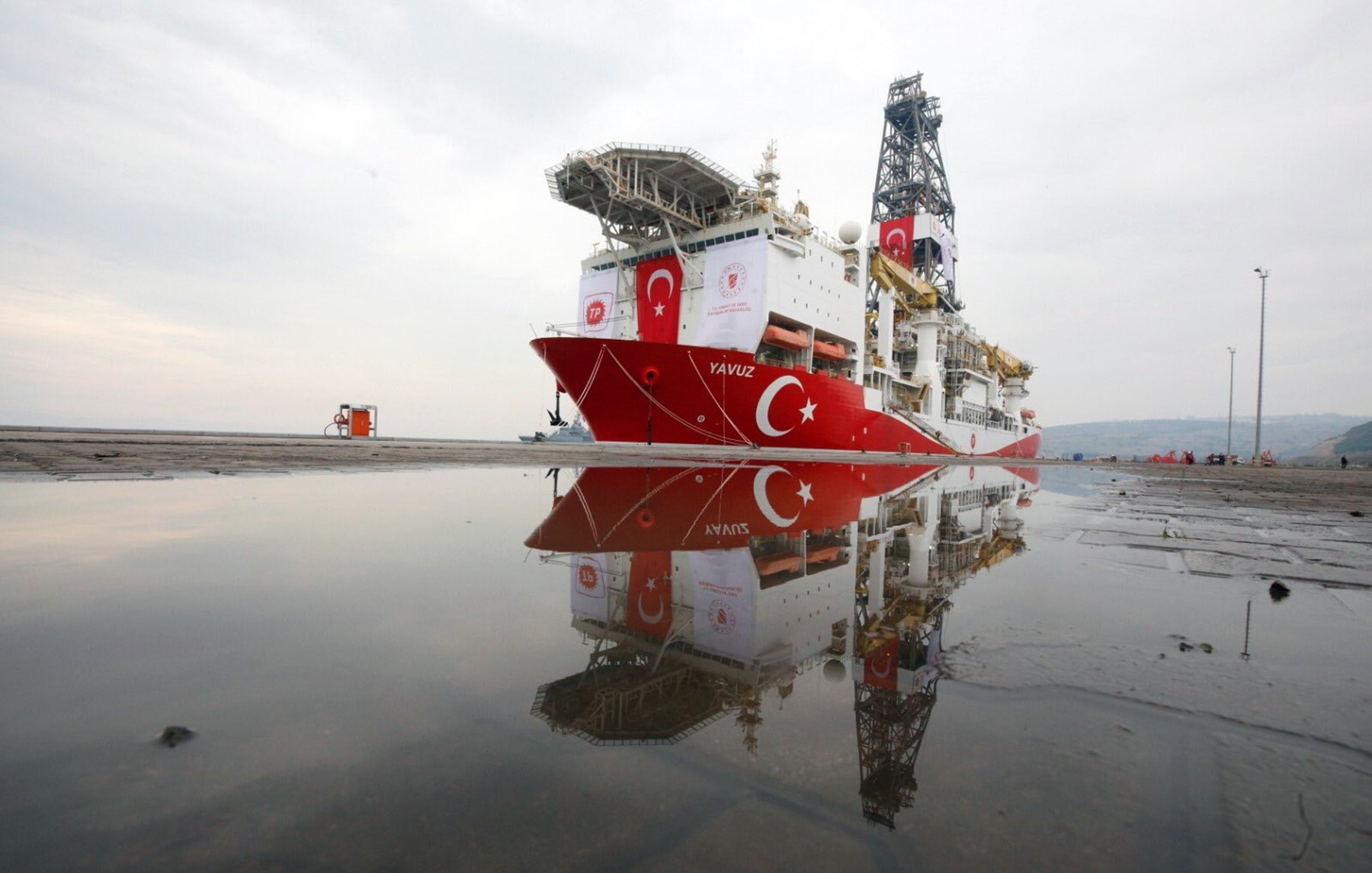 L'image montre un navire de forage maritime, nommé "Yavuz", qui est amarré au port. Le bateau arbore des couleurs dominantes de rouge et blanc, représentant le drapeau turc. On peut également voir des équipements de forage sur le pont du navire. Dans la partie inférieure de l'image, il y a des réflexions du navire dans une flaque d'eau sur le sol. Le ciel en arrière-plan est nuageux, créant une atmosphère calme.