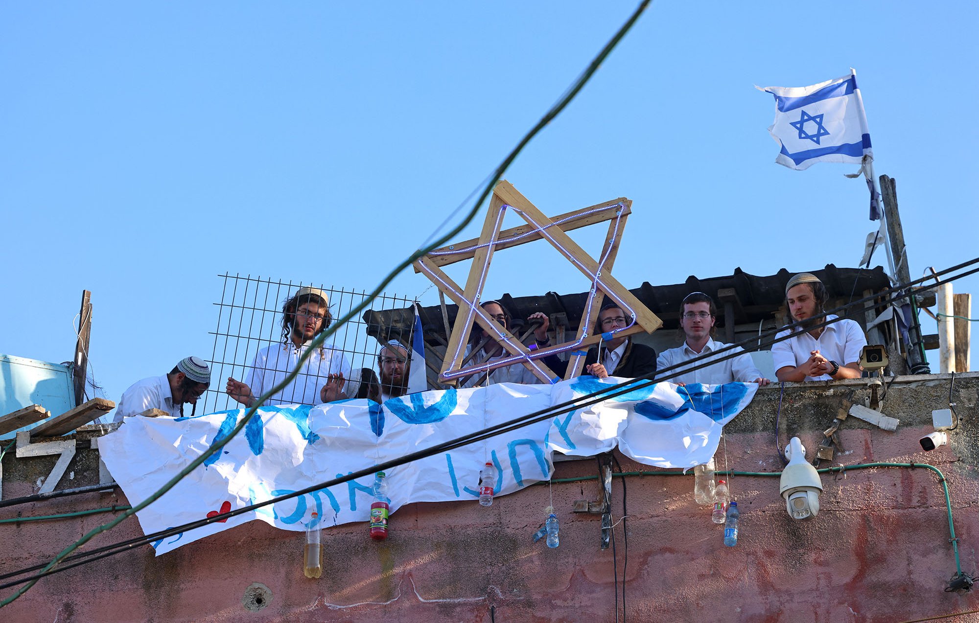 L'image montre un groupe de personnes rassemblées sur un toit. Elles portent des vêtements traditionnels, et certaines tiennent un symbole en forme d'étoile de David. En arrière-plan, il y a un drapeau israélien flottant. On peut également voir une banderole accrochée, suggérant un message ou une revendication. Le ciel est dégagé, et il y a une ambiance de rassemblement communautaire.