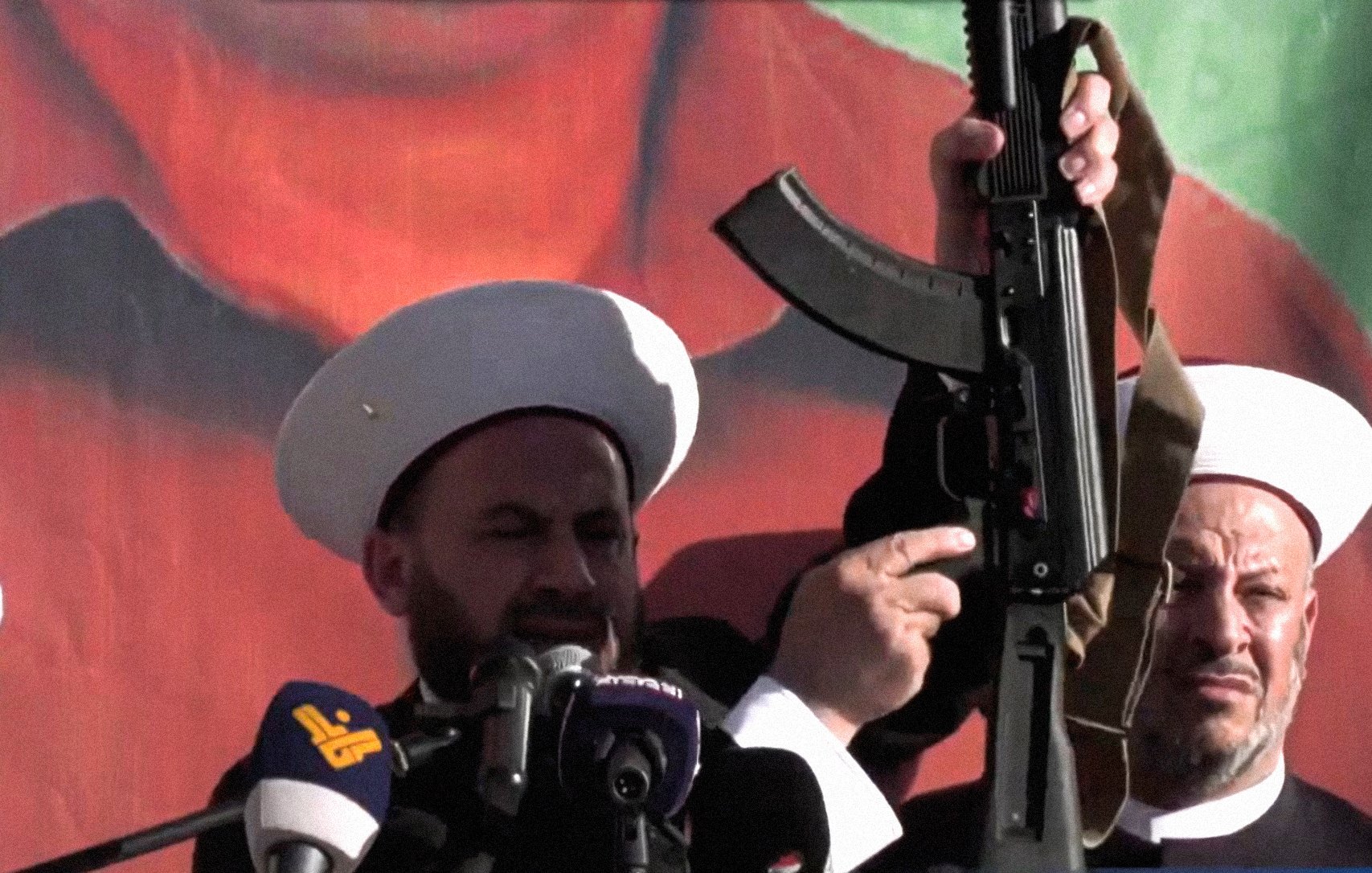 L'image montre deux hommes portant des chapeaux traditionnels, se tenant devant un grand panneau ou une affiche représentant une figure politique ou historique. L'un des hommes brandit une arme, probablement un fusil d'assaut, en faisant un geste qui attire l'attention du public. L'atmosphère semble solennelle et les deux hommes semblent s'adresser à une foule, ce qui suggère un contexte d'engagement politique ou religieux.