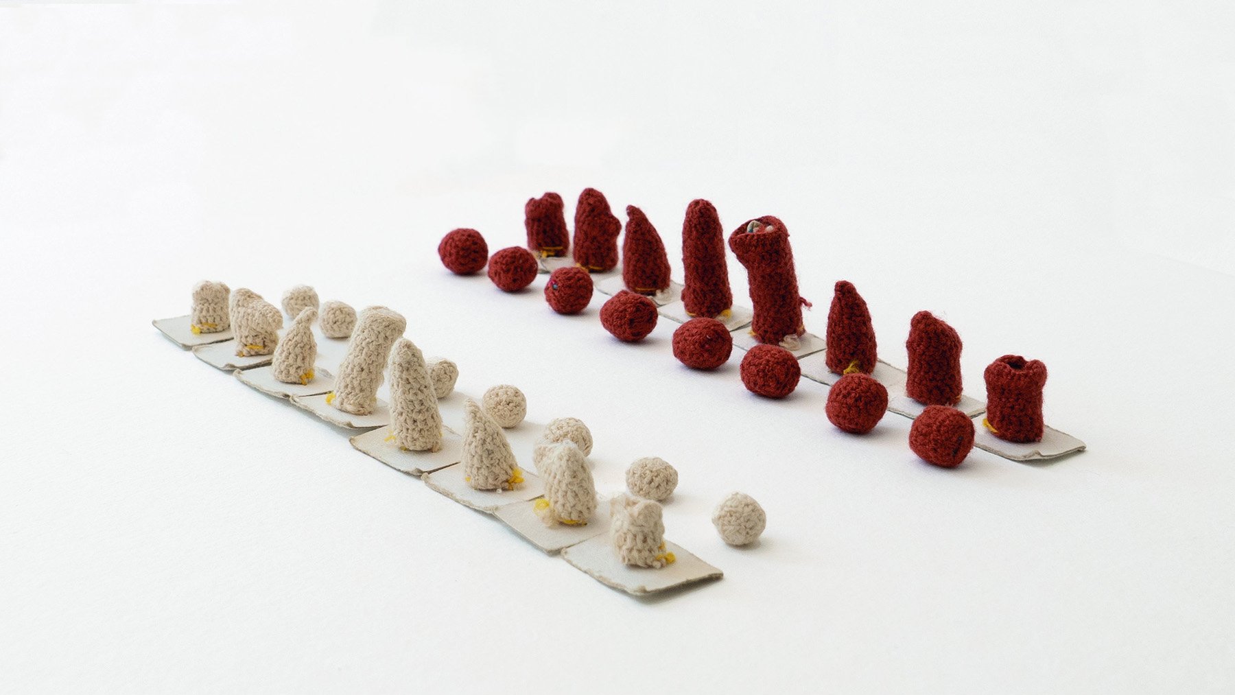 L'image montre une composition artistique composée de petits objets en crochet. Il y a deux groupes disposés sur une base claire : l'un en rouge et l'autre en beige. Les objets de chaque côté varient en forme et en taille, avec des cônes et des boules. La présentation est minimaliste, mettant en avant les textures et les couleurs des créations. L'arrangement est symétrique, créant un effet visuel intéressant.