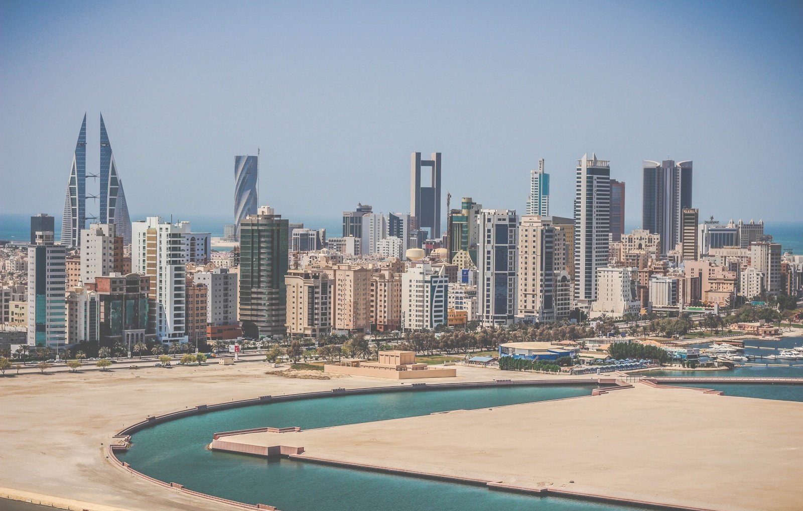 L'image montre un paysage urbain moderne avec une vue sur une ville côtière. On peut voir de nombreux immeubles de grande hauteur, représentant une architecture contemporaine, avec des designs variés allant des structures angulaires aux façades en verre. À l'arrière-plan, la mer est visible, ainsi qu'une baie ou un port. Le ciel est clair et ensoleillé, ce qui donne une impression de chaleur et de vivacité à la scène. La composition évoque une ambiance urbaine dynamique, typique de villes en développement.