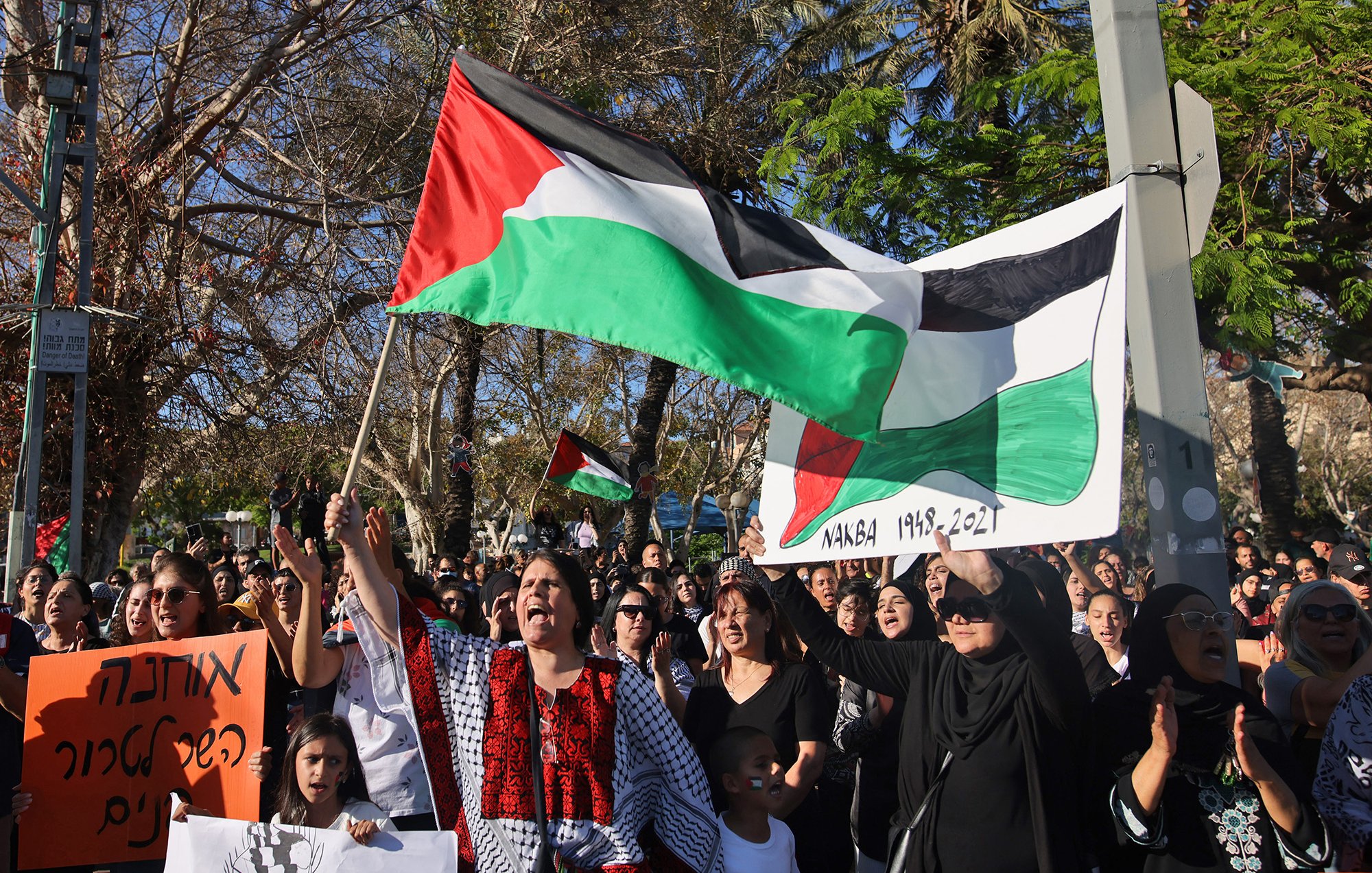 L'image montre une manifestation où des personnes tiennent des drapeaux palestiniens et des pancartes. La foule semble rassemblée pour exprimer une opinion ou un soutien, avec des femmes et des enfants présents. L'arrière-plan inclut des arbres et un environnement urbain. Les participants portent des vêtements variés, certains ayant des motifs traditionnels. L'atmosphère paraît chargée d'émotion et de solidarité.