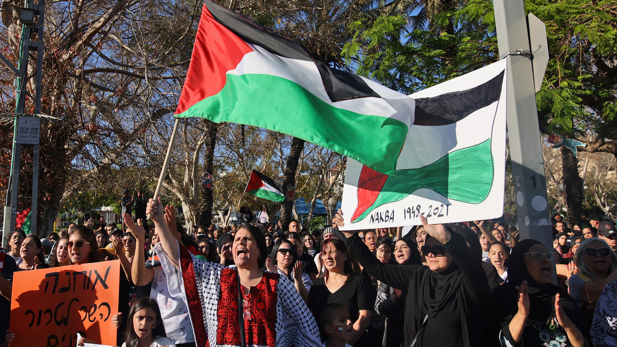 L'image montre une manifestation où des personnes tiennent des drapeaux palestiniens et des pancartes. La foule semble rassemblée pour exprimer une opinion ou un soutien, avec des femmes et des enfants présents. L'arrière-plan inclut des arbres et un environnement urbain. Les participants portent des vêtements variés, certains ayant des motifs traditionnels. L'atmosphère paraît chargée d'émotion et de solidarité.