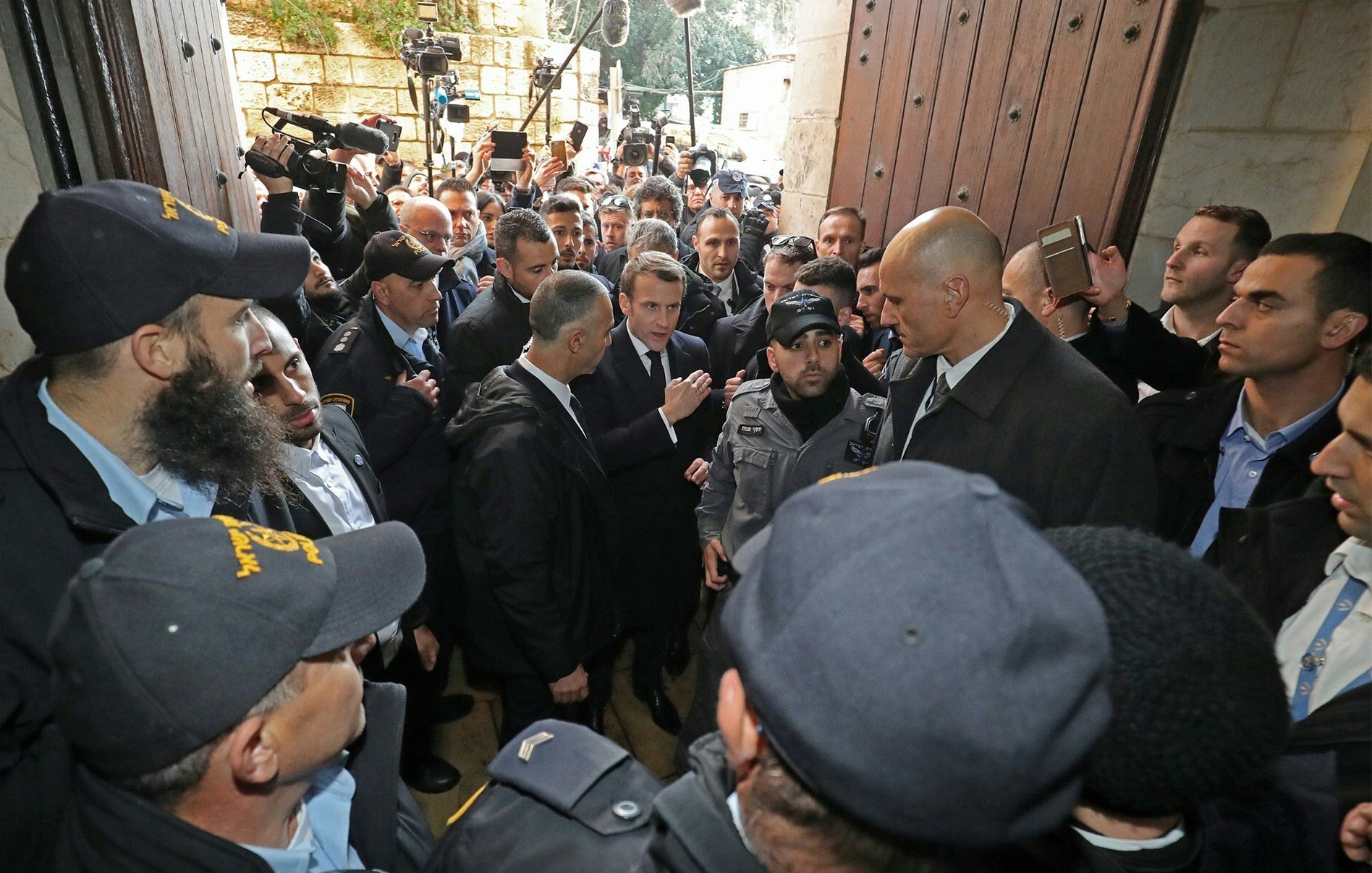 L'image montre une scène animée avec de nombreuses personnes rassemblées autour d'un groupe central. Des agents de sécurité, en uniforme, semblent encadrer des individus, probablement des personnalités importantes. L'atmosphère est tendue, avec des caméras et des journalistes présents, suggérant que cet événement pourrait avoir une importance médiatique. L'ensemble se déroule devant une grande porte, indiquant un lieu significatif, possiblement un bâtiment officiel ou historique.