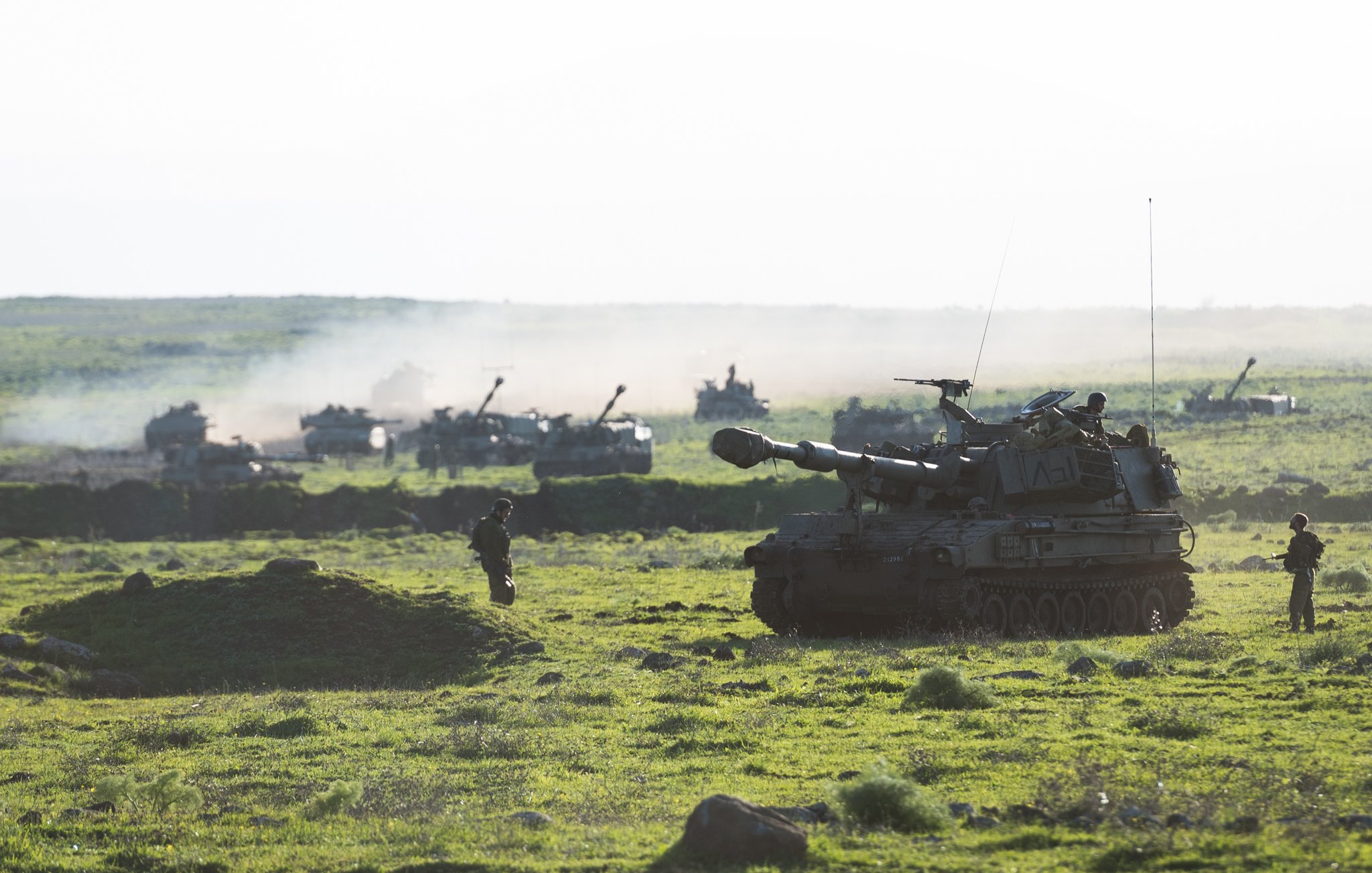 L'image montre un paysage militaire verdoyant avec des chars d'assaut et d'autres véhicules en arrière-plan. On peut voir un char principal en premier plan, déployé sur un terrain herbeux, tandis que plusieurs autres véhicules sont visibles plus loin, entourés d'une légère brume ou fumée. Quelques soldats sont présents, donnant une impression d'activité militaire dans un environnement naturel. La lumière est douce, suggérant peut-être un moment du jour comme le lever ou le coucher du soleil.