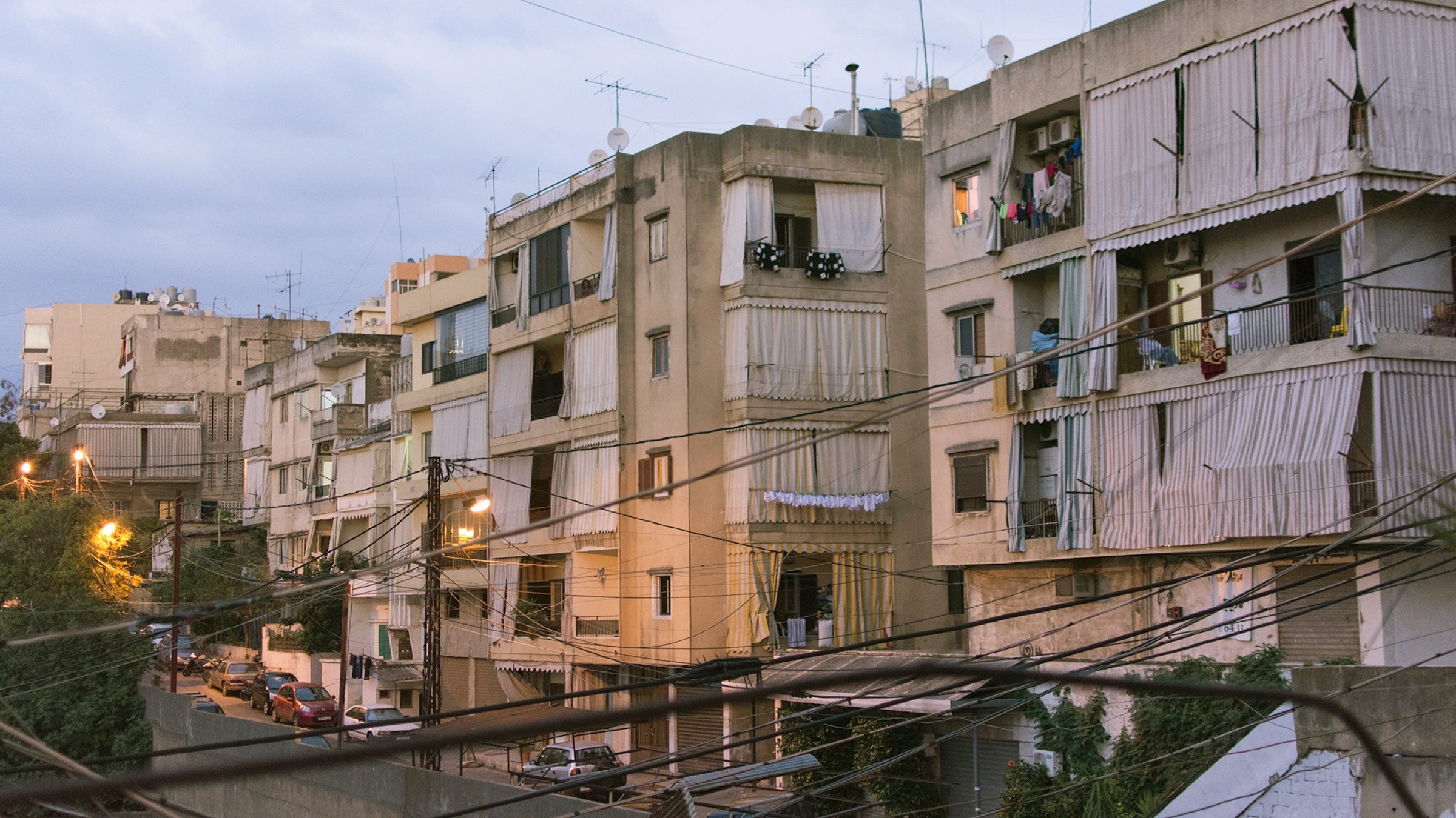 L'image montre un ensemble d'immeubles résidentiels dans une zone urbaine. Les bâtiments semblent anciens et sont de plusieurs étages, avec des balcons où l'on peut voir des vêtements étendus. La lumière du soir crée une atmosphère douce, et des fils électriques traversent l'image. On aperçoit également des voitures garées le long de la rue en contrebas. L'ensemble donne une impression de vie citadine typique, avec un mélange de modernité et de caractère ancien.