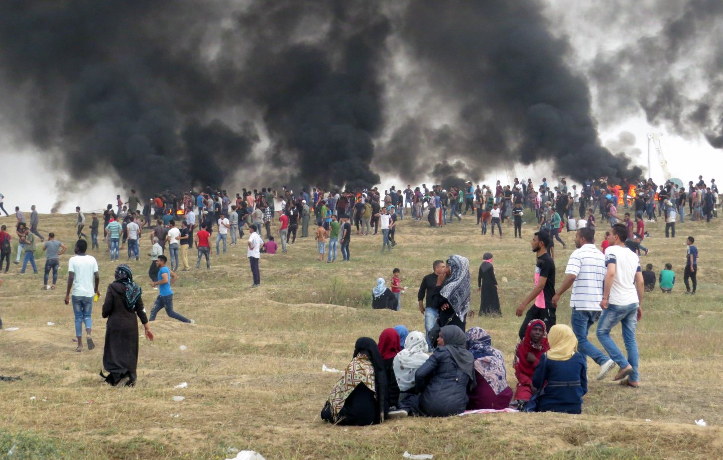 L'image montre une grande foule de personnes rassemblées sur une colline, avec d'importants nuages de fumée noire émanant en arrière-plan. Les gens semblent se tenir debout ou marcher, certains regardant vers la fumée. Au premier plan, un groupe de personnes est assis sur le sol, tandis que d'autres se déplacent autour d'eux. L'ambiance est tendue, indiquant une situation d'agitation ou de protestation. Le paysage est principalement sec avec quelques herbes.