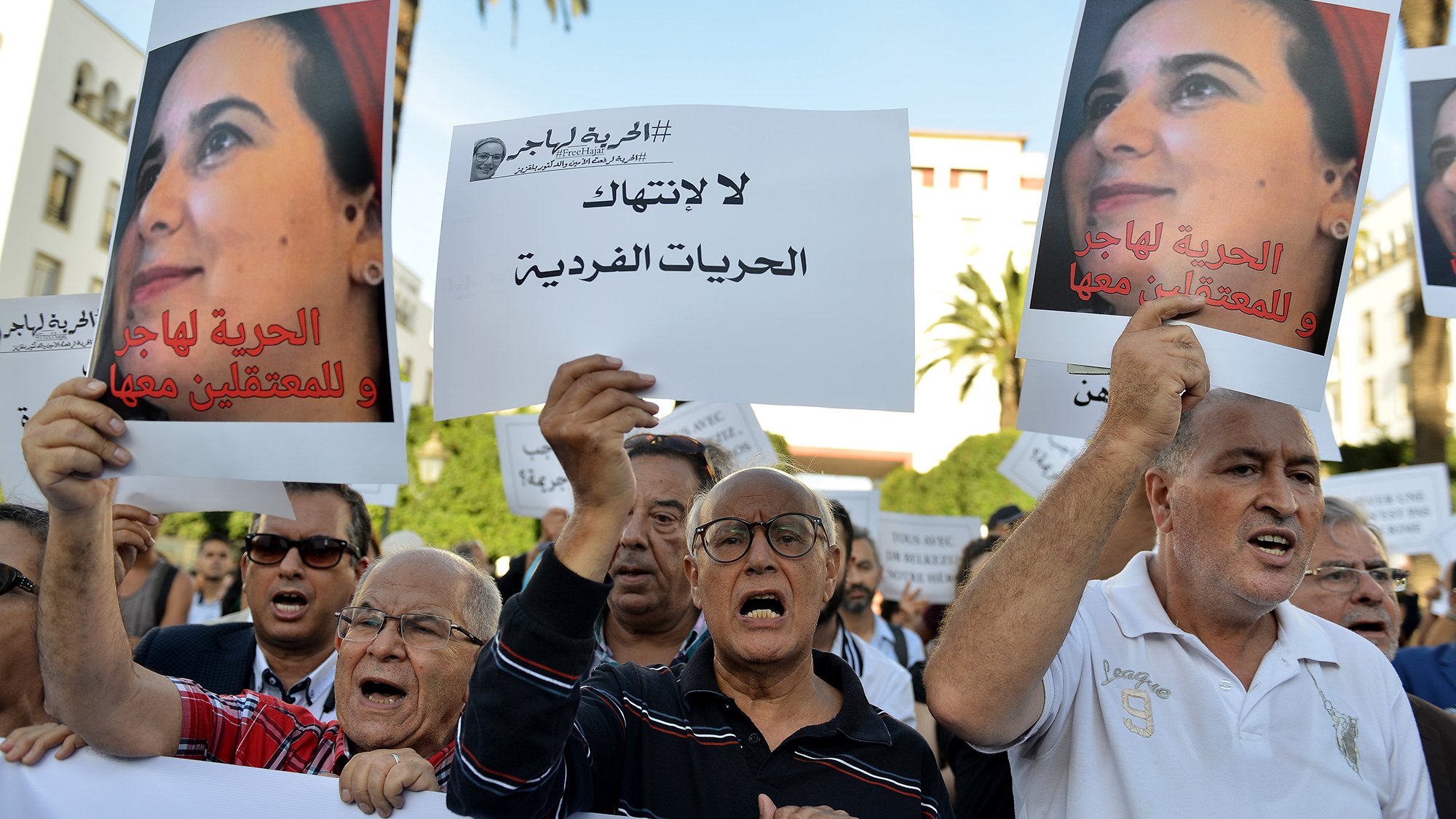 L'image montre une manifestation avec un groupe de personnes tenant des pancartes. Les manifestants semblent exprimer leur désaccord et revendiquer des droits, notamment en ce qui concerne les libertés individuelles. Certains affichent des expressions de colère ou de détermination, tandis que d'autres portent des signes clairs de solidarité. En arrière-plan, on peut voir des palmiers et un bâtiment, suggérant un cadre urbain. L'atmosphère est engagée et militante, reflétant une lutte pour des causes sociales ou politiques.