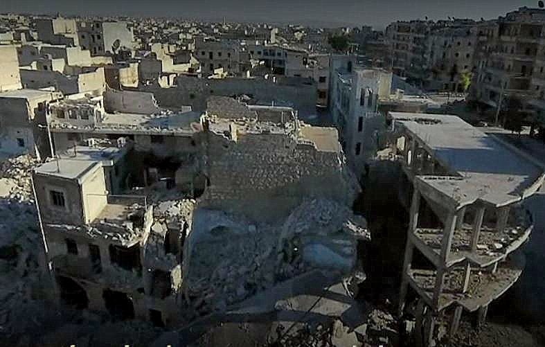 L'image montre un paysage urbain dévasté, probablement dans une zone de conflit. On y voit des bâtiments en ruines, certains complètement effondrés, d'autres partiellement intacts. Le sol est jonché de débris, témoignant des destructions causées par des bombardements ou des combats. L'environnement paraît déserté, avec peu ou pas de signes de vie, illustrant la tragédie et les conséquences dramatiques de la guerre sur les infrastructures urbaines.