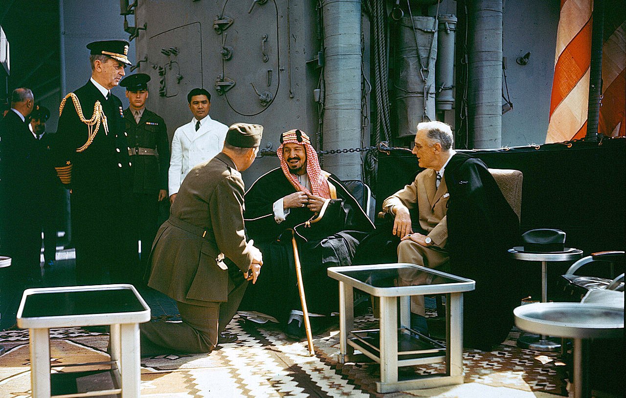 L'image montre un groupe de personnalités en discussion sur un navire. Au centre, un homme en tenue traditionnelle semble rire ou sourire, assis avec une canne. À sa gauche, un homme en habit sombre, tandis qu'un autre, en uniforme militaire, est à genoux devant eux. En arrière-plan, d'autres personnes observent la scène, dont un officier en uniforme. Le décor est richement décoré avec des tapis et des éléments nautiques.