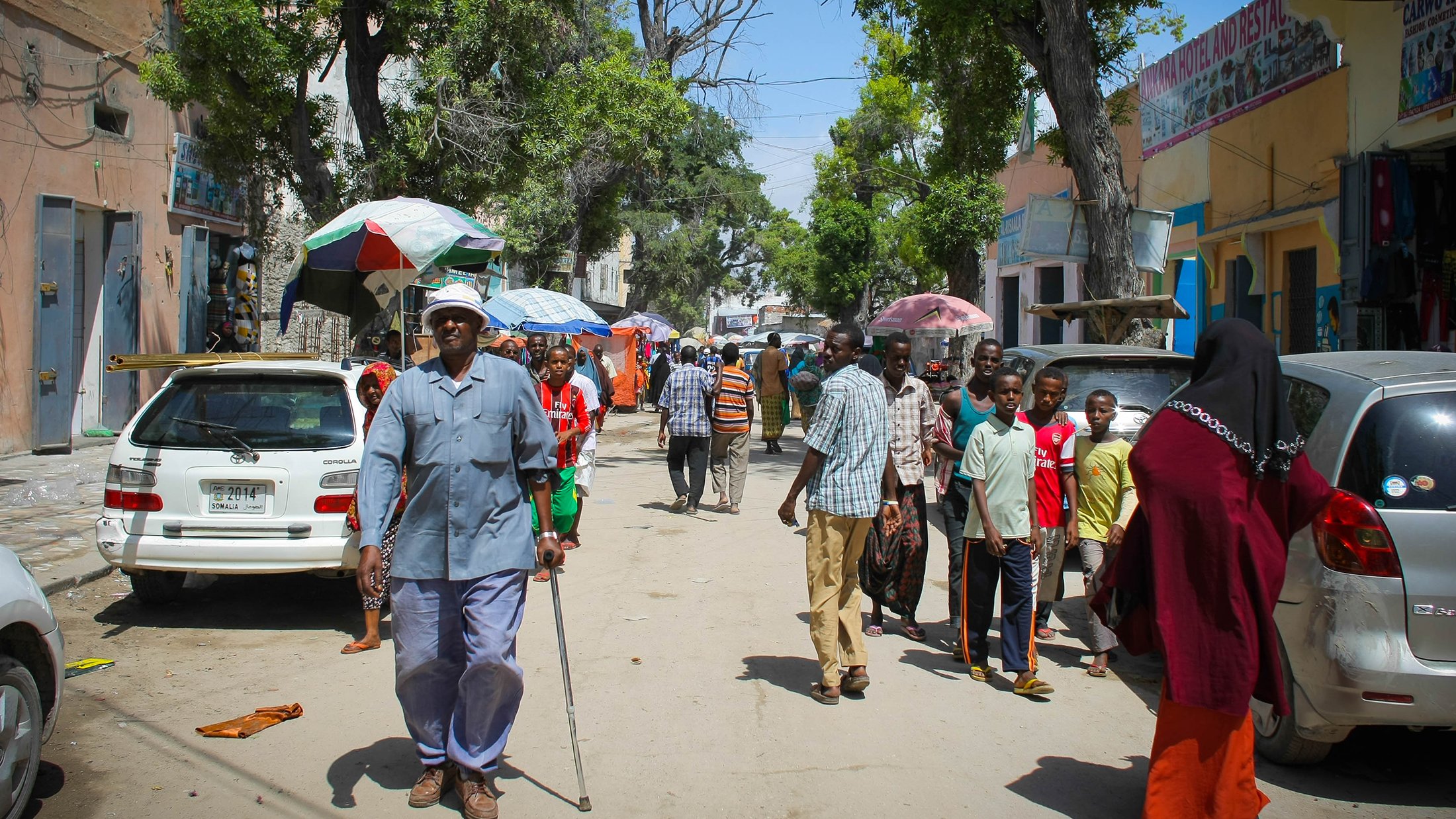 L'image montre une rue animée avec des groupes de personnes marchant. On peut voir un homme âgé utilisant une canne, habillé en bleu, qui semble avancer lentement. Plusieurs personnes sont dispersées le long de la rue, certaines se trouvent près de voitures garées. Des ombrelles colorées sont visibles, probablement pour se protéger du soleil. Les bâtiments en arrière-plan semblent être de petits commerces ou habitations. L'ambiance générale dénote une vie quotidienne dynamique dans un environnement urbain.