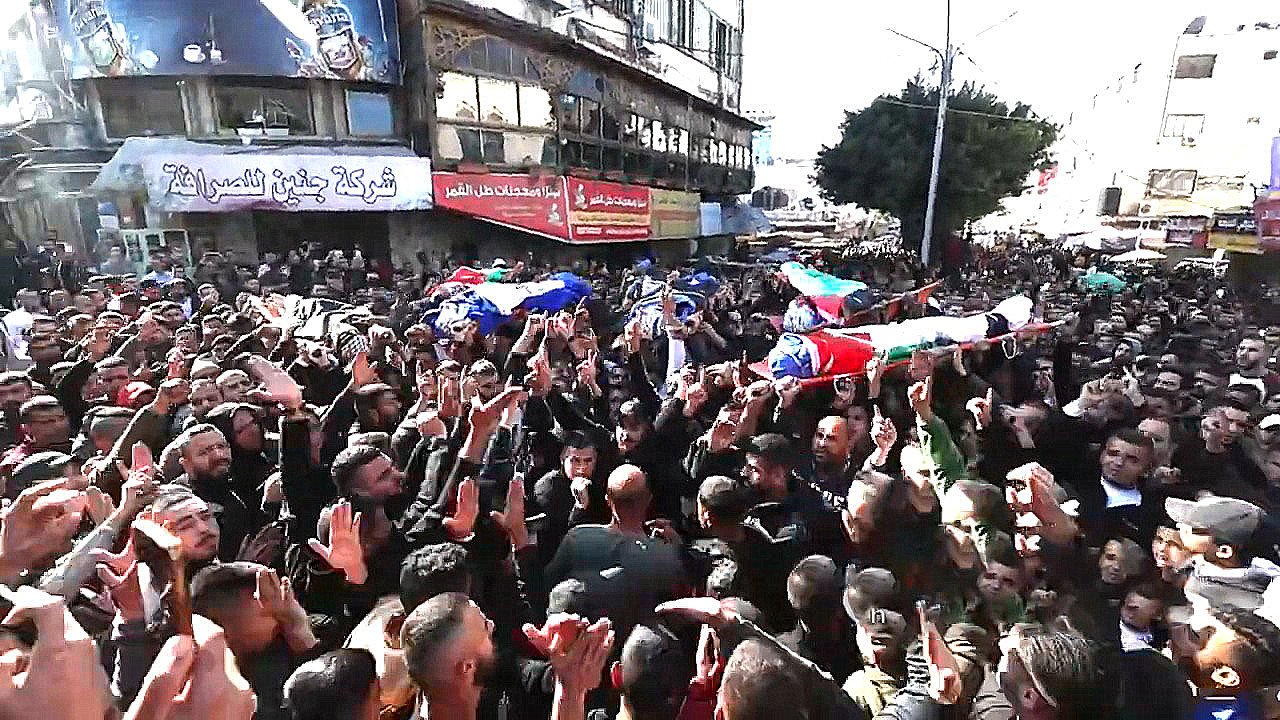 L'image montre une grande foule rassemblée dans une rue, avec des personnes portant des drapeaux et des pancartes. Il y a des cercueils sur des supports, symbolisant un hommage ou des funérailles. La foule est visiblement très engagée, levant les mains et exprimant des émotions fortes. L'environnement urbain est marqué par des bâtiments et des affiches en arrière-plan. L'atmosphère est chargée de gravité et de solidarité.