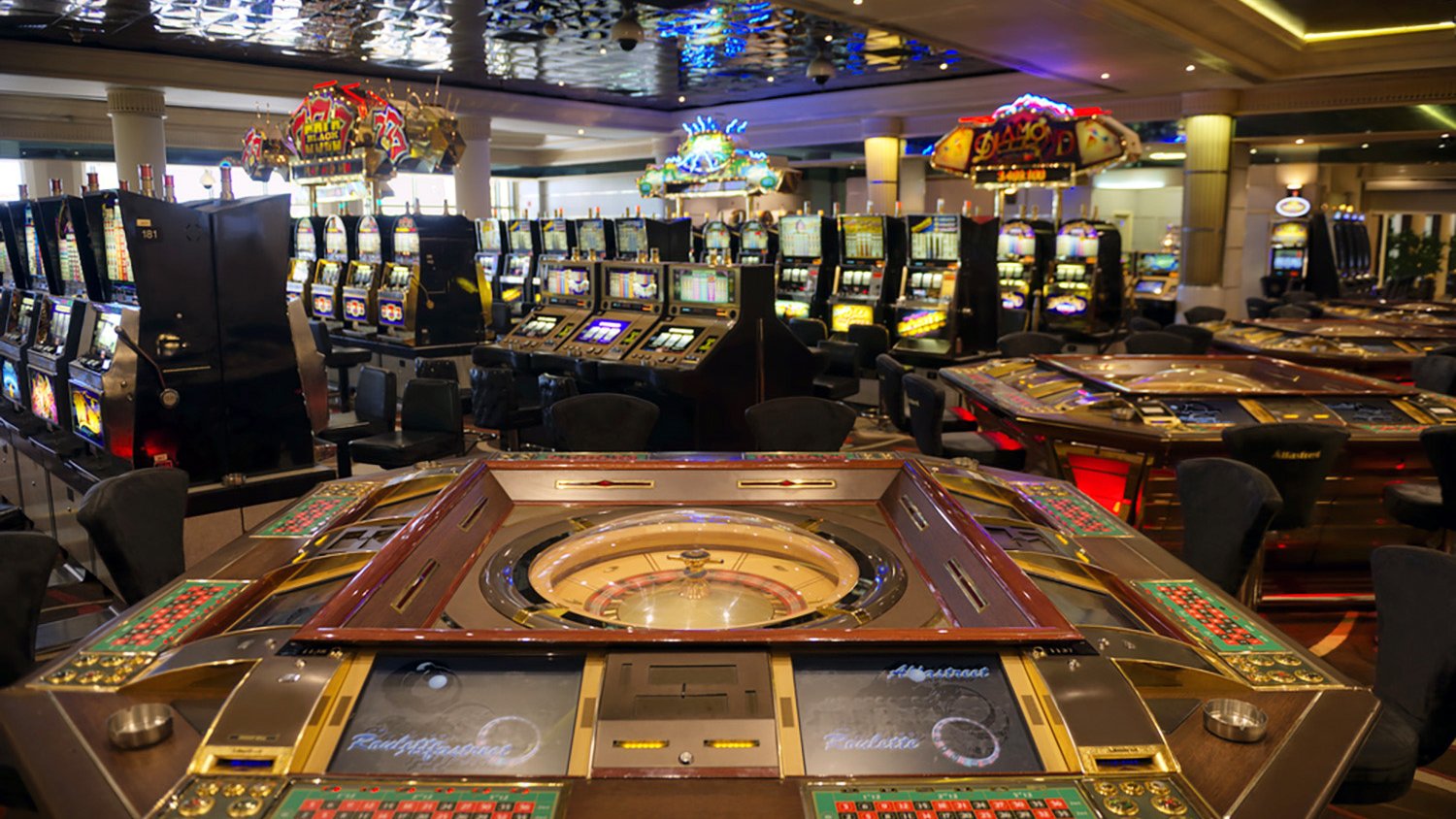 L'image montre l'intérieur d'un casino, avec plusieurs tables de jeux et des machines à sous. Au premier plan, on peut voir une grande table de roulette, entourée de plusieurs sièges. À l'arrière-plan, on distingue de nombreuses machines à sous illuminées, créant une atmosphère animée et colorée. Le plafond est décoré, ajoutant à l'élégance de l'espace. L'ensemble dégage une ambiance de divertissement et de luxe.