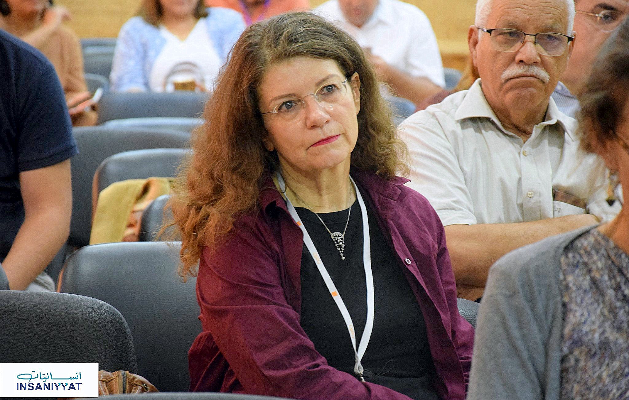 L'image montre une femme assise dans une salle de conférence ou de réunion, avec une expression pensive. Elle porte un haut de couleur sombre et a de longs cheveux bouclés. En arrière-plan, on peut voir d'autres personnes dans une atmosphère assez calme. La femme semble concentrée sur ce qui se passe autour d'elle.