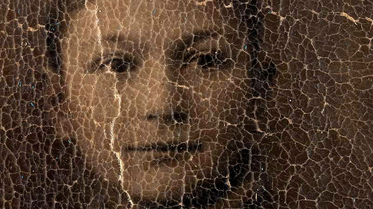 L'image représente un portrait d'une personne, probablement un jeune garçon, dont les traits sont légèrement obscurcis par un certain vieillissement et des fissures à la surface. Le visage montre des expressions délicates et le regard engageant, mais les détails sont atténués à cause de l'état usé de la photographie. Le fond semble sombre, ce qui accentue l'apparence vieillie de l'image.