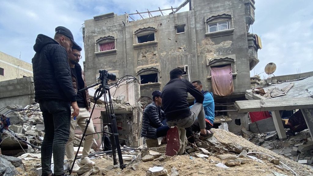 L'image montre un groupe de personnes se tenant sur des décombres, probablement après un effondrement de bâtiment. Ils semblent être en train de filmer ou de prendre des photos, avec du matériel de caméra visible. En arrière-plan, on peut voir une structure endommagée, avec des fenêtres brisées et des pans de mur manquants. Le ciel est nuageux, ce qui ajoute à l'atmosphère sombre de la scène. L'environnement est chaotique, reflétant les conséquences d'une destruction.