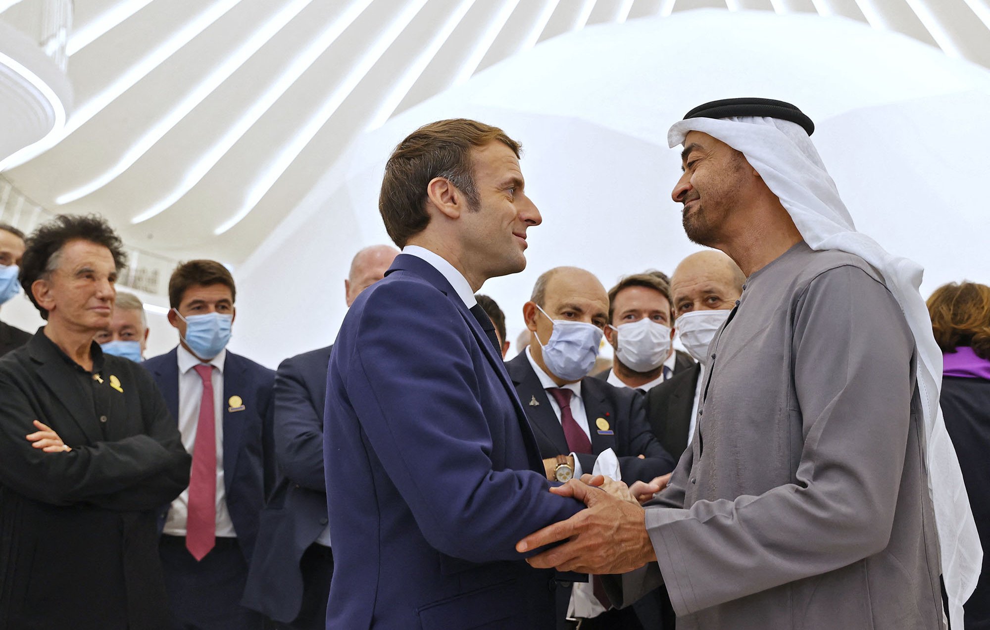 L'image montre une rencontre entre deux dirigeants, avec l'un portant un costume et l'autre vêtu d'une tenue traditionnelle des Émirats. Ils semblent engager une conversation amicale, se serrant la main avec des expressions de cordialité. En arrière-plan, plusieurs personnes les regardent, certaines portant des masques. L'environnement est moderne et lumineux, avec des éléments architecturaux contemporains.