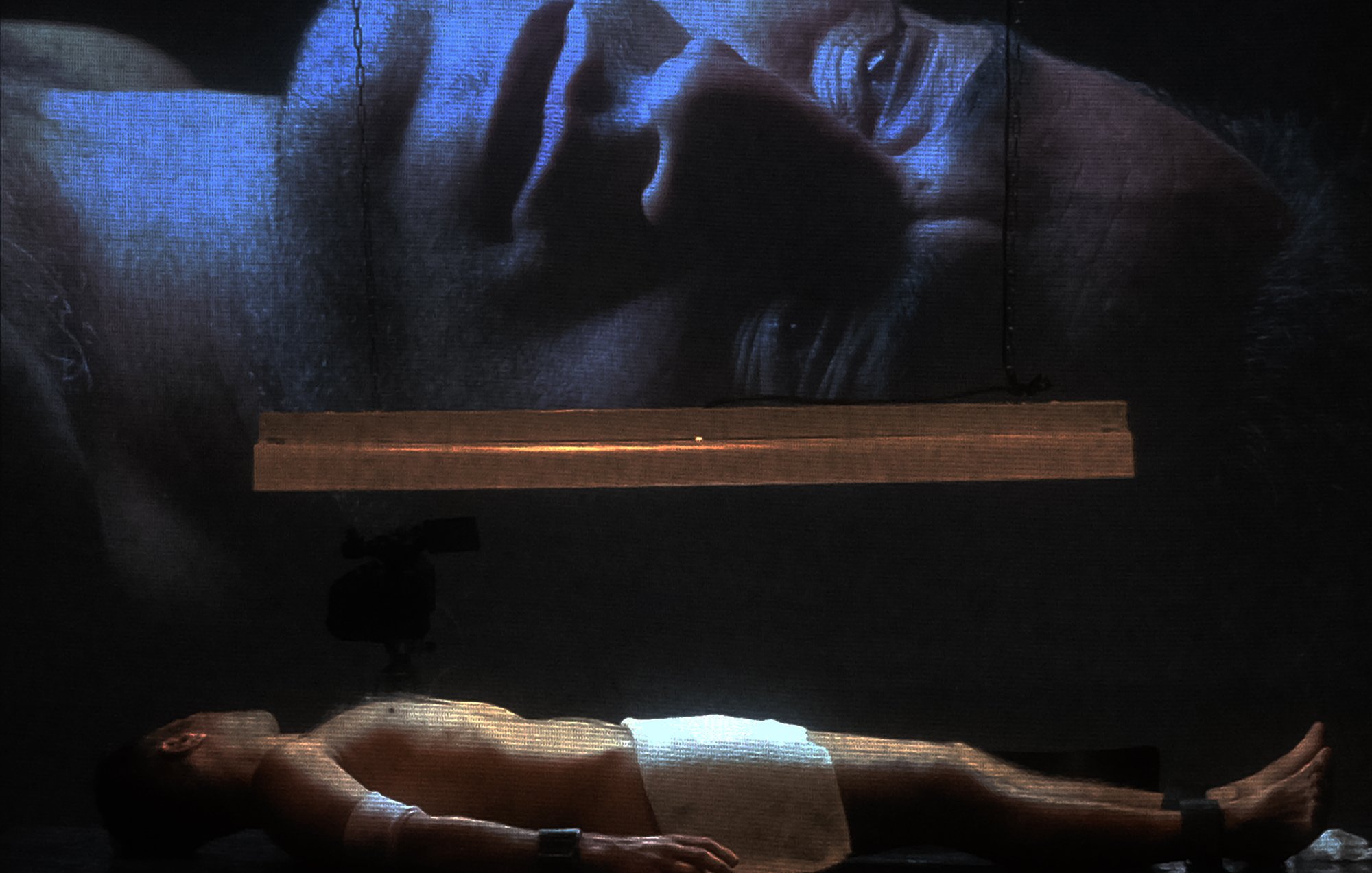 L'image montre un homme allongé sur une surface, partiellement nu, avec une serviette couvrant son corps. Au-dessus de lui, une projection de son visage est affichée sur un grand écran, créant un effet visuel puissant. La lumière provient d'une source suspendue, ajoutant une atmosphère dramatique à la scène. Les détails du visage projeté sont clairement visibles, tandis que l'homme au sol semble dans un état de vulnérabilité. L'ensemble évoque des thèmes de réflexion, d'introspection ou de méditation.