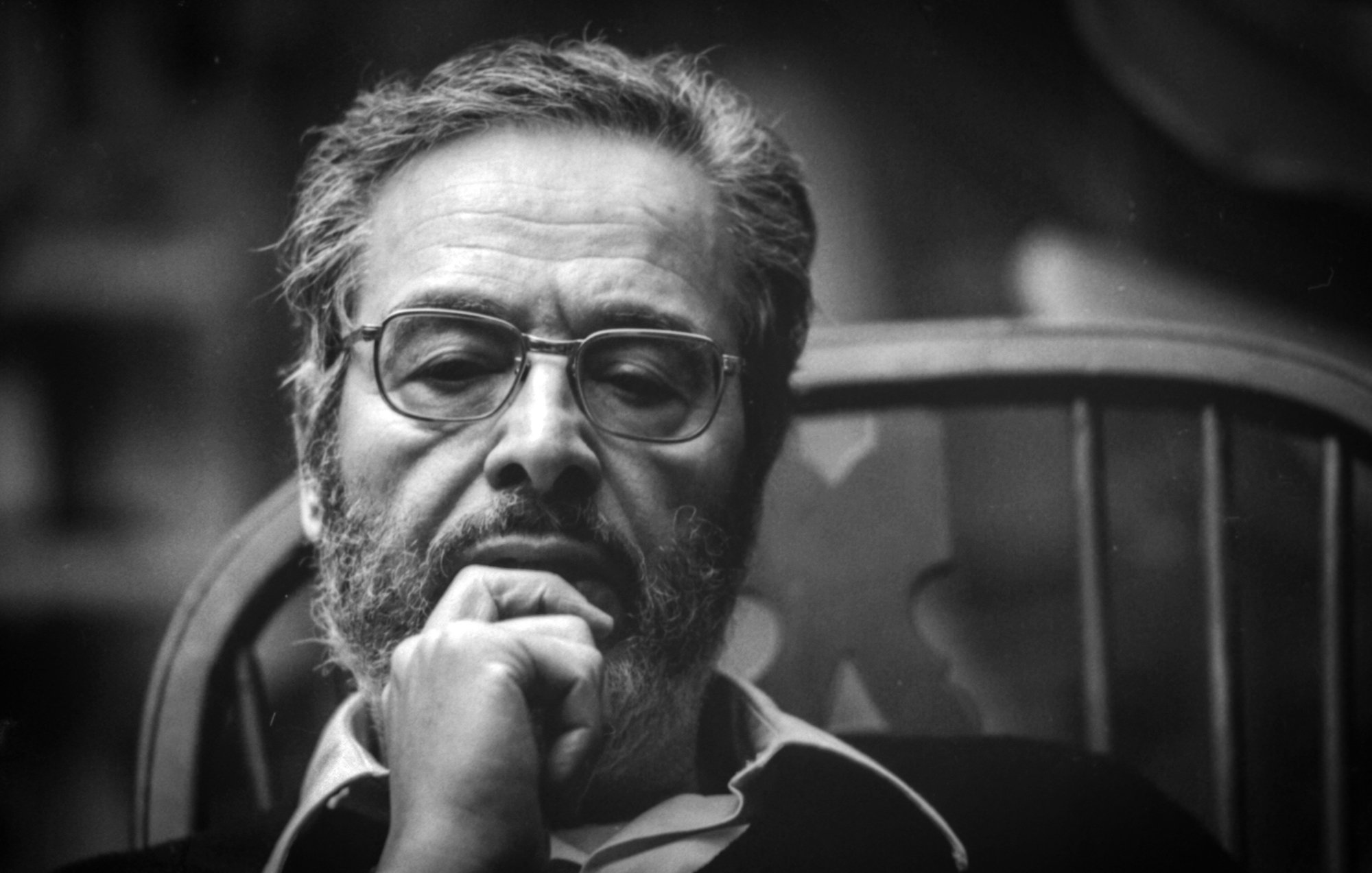 L'image montre un homme assis sur une chaise, arborant une barbe et des lunettes. Il semble profond dans ses pensées, avec une main sous le menton. L'éclairage donne un aspect sérieux à son expression, et le fond flou met en avant son visage. L'atmosphère est introspective et contemplative.