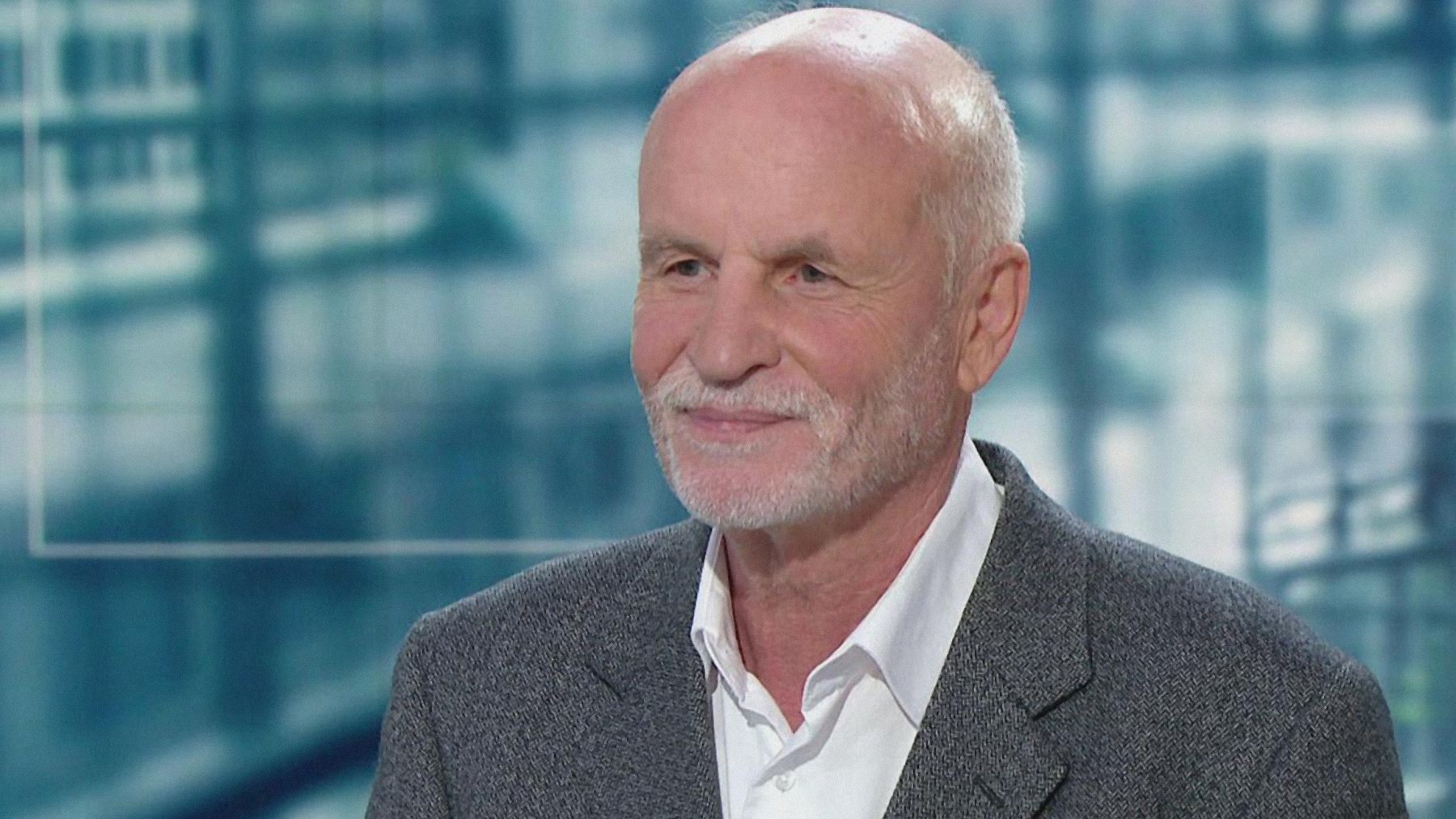 Homme âgé avec cheveux gris, souriant, portant un costume sobre sur fond flou.
