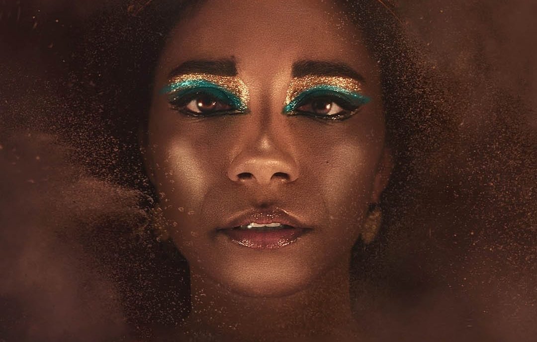 L'image montre une personne au visage ornée, avec un maquillage très artistique mettant en avant des yeux accentués par une ombre à paupières bleu vif. La personne porte une coiffe dorée en forme de serpents, ajoutant à l'aspect majestueux de l'ensemble. Le fond est flou et semble composé de particules dorées, créant une atmosphère mystique et intrigante. L'expression du visage est à la fois sereine et mystérieuse.