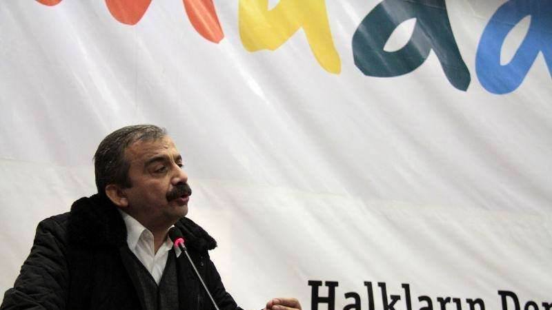 L'image montre un homme parlant lors d'un événement ou d'une conférence. Il est vêtu d'un manteau sombre et d'une chemise blanche. Le fond affiche une grande bannière avec des mots colorés, dont "Halkların Derneği", qui signifie "L'Association des Peuples" en turc. L'homme semble engagé dans son discours, utilisant des gestes pour illustrer ses propos.