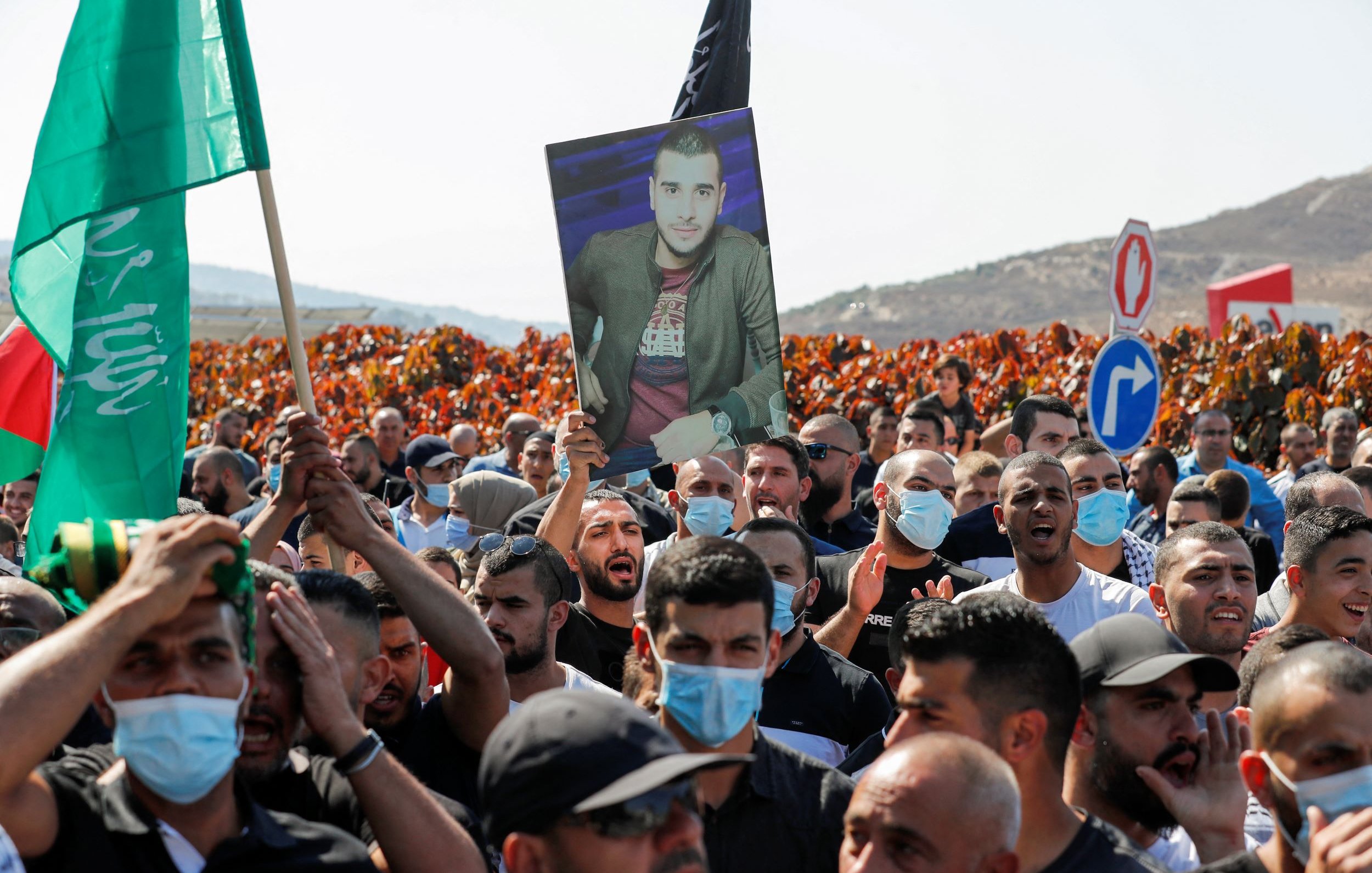 L'image montre une manifestation où une foule dense est présente. Au premier plan, des manifestants tiennent des drapeaux, dont un vert, et certains agitent des pancartes. Parmi eux, une personne porte un grand portrait d'un individu, visiblement en hommage ou en soutien. Les manifestants semblent engagés et déterminés, et ils portent tous des masques sanitaires, indiquant un contexte de pandémie. En arrière-plan, des collines se dessinent, avec des panneaux de signalisation visibles. L'atmosphère semble chargée d'émotion et de revendications.