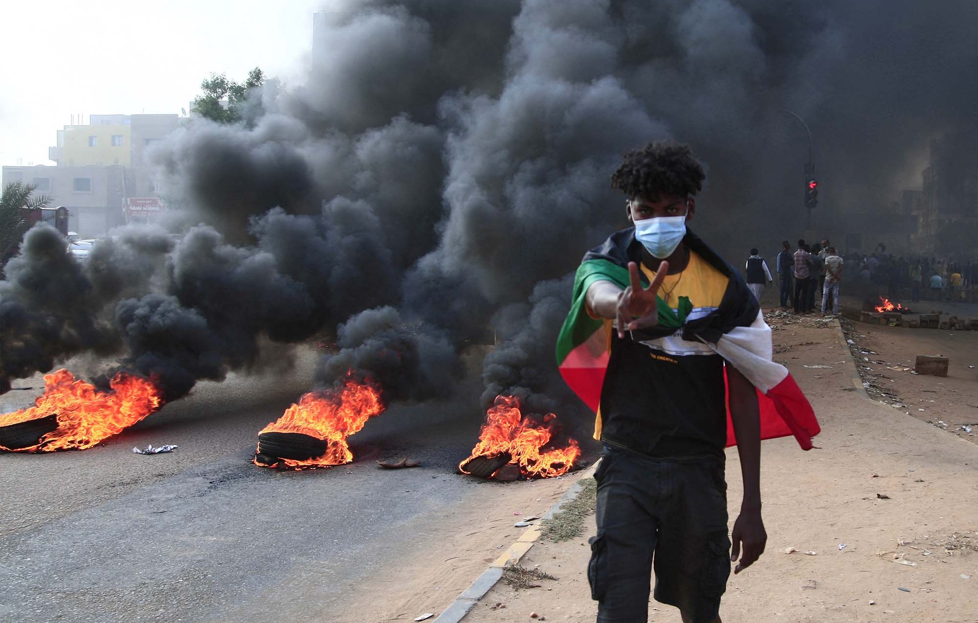 L'image montre un jeune homme marchant sur une route pendant une manifestation. Il porte un masque et fait un signe de paix avec sa main. En arrière-plan, on peut voir des pneus enflammés et une épaisse colonne de fumée noire s'élevant dans l'air. La scène dégage une impression de tension et de protestation, et il y a un groupe de personnes visibles derrière lui.
