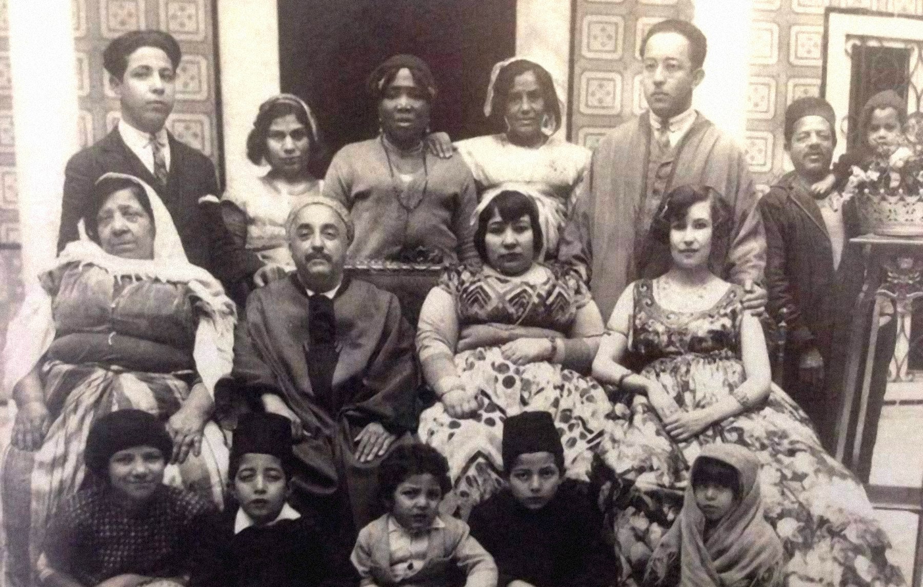 L'image présente un groupe de personnes, probablement une famille ou une communauté, posant ensemble dans un décor intérieur. On peut voir des hommes et des femmes de différentes générations, ainsi que des enfants, vêtus de tenues traditionnelles. L'atmosphère semble conviviale et chaleureuse. L'arrière-plan est orné de motifs, ajoutant une touche historique à la scène.