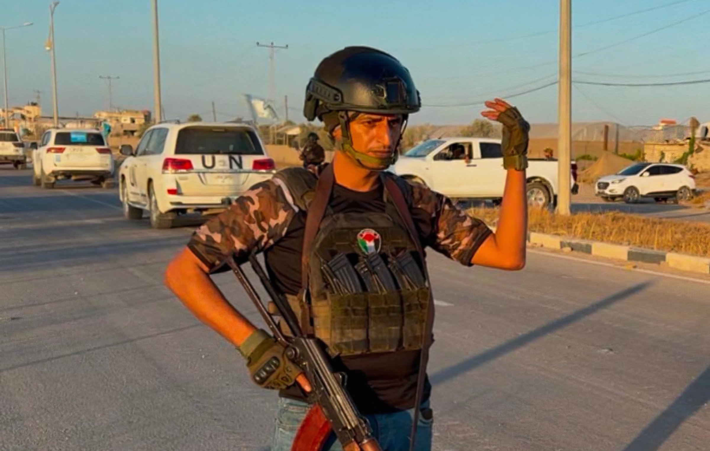 Un homme armé, en tenue militaire, se tient sur une route avec des voitures en arrière-plan.