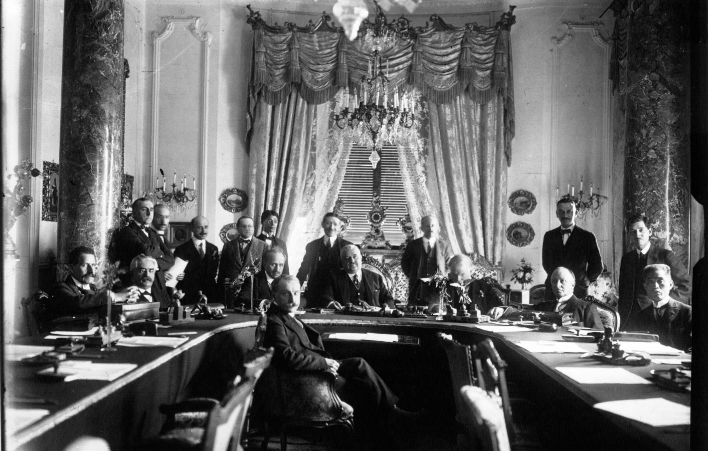 L'image montre une grande réunion dans une salle ornée. Au centre, plusieurs hommes sont assis autour d'une grande table, tandis que d'autres se tiennent debout en arrière-plan. La pièce est décorée avec des rideaux élégants et des éléments architecturaux raffinés, tels que de grands lustres et des colonnes. Les participants semblent engagés dans une discussion sérieuse, reflétant l'importance de l'événement. L'atmosphère est formelle et historique.