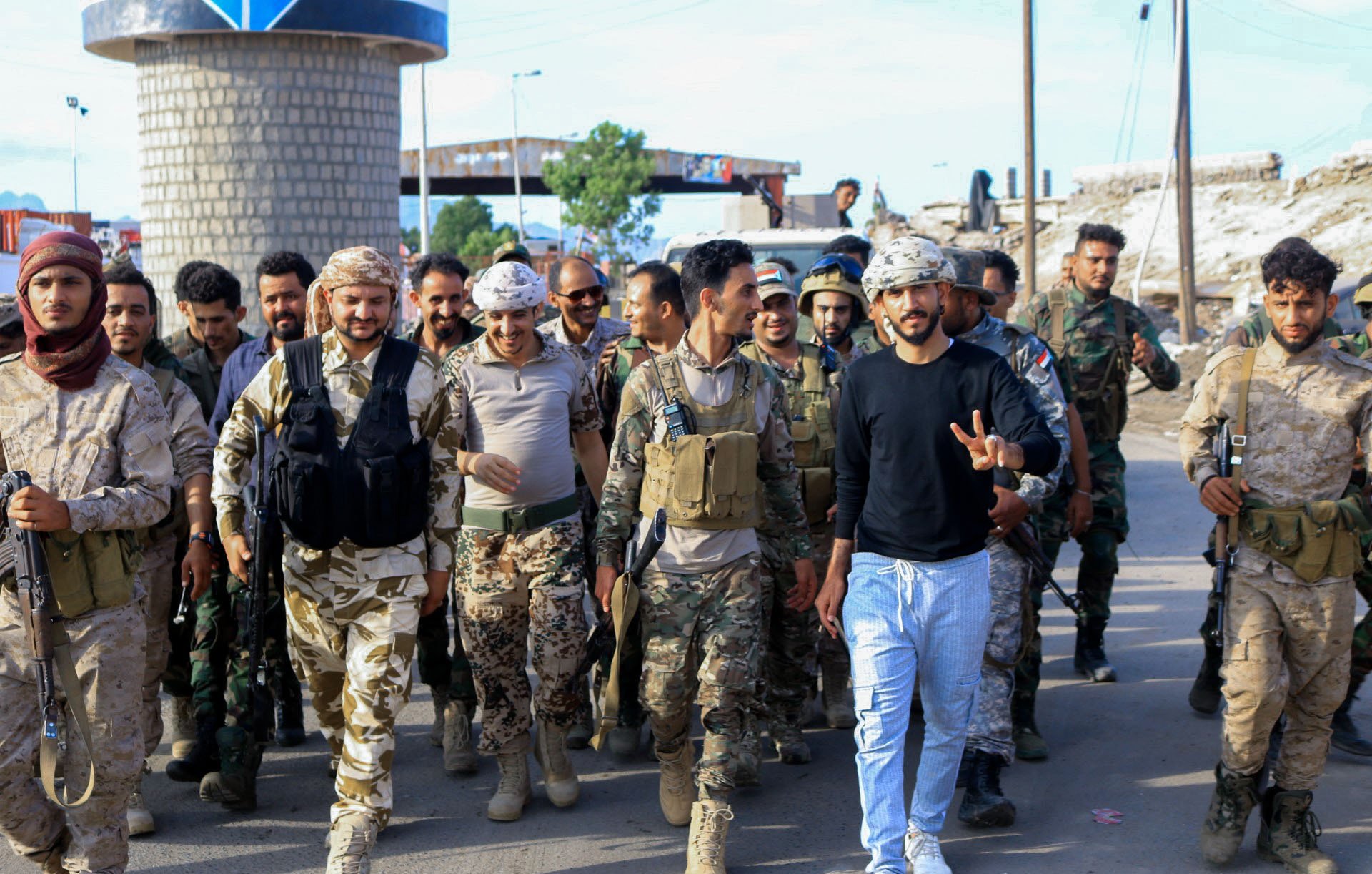 L'image montre un groupe de soldats marchant ensemble dans une zone urbaine. Parmi eux, on peut voir des militaires en uniformes camouflés et d'autres en tenues militaires variées. Certains portent des gilets tactiques et des casques. On remarque une atmosphère de camaraderie et de détermination parmi les membres du groupe, avec une personne au centre faisant un signe de paix. En arrière-plan, des infrastructures urbaines sont visibles, ce qui indique un environnement de mission militaire.