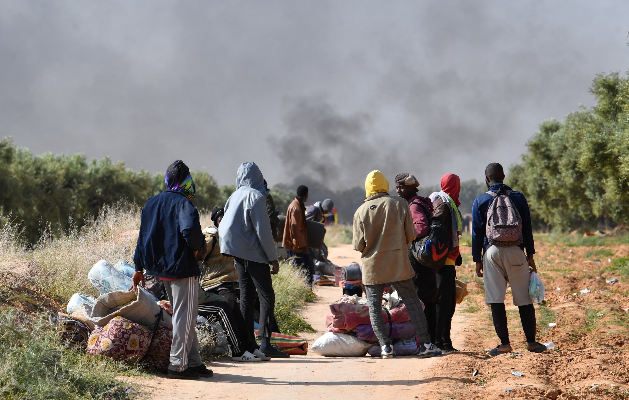 Un groupe de personnes se tient sur un chemin, entouré de fumée et de bagages.