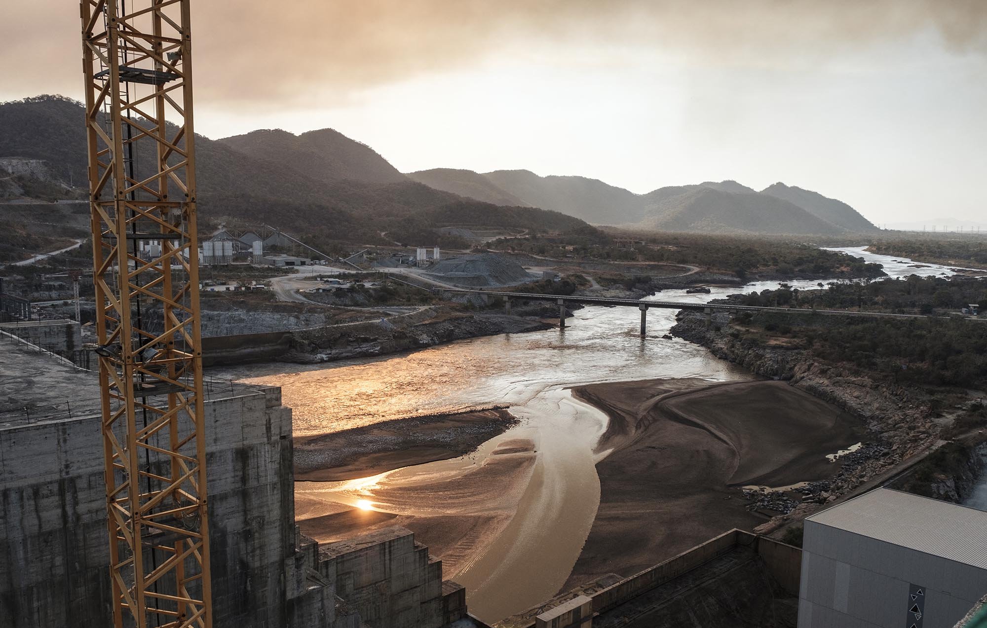 L'image montre un paysage industriel au bord d'une rivière. On y voit des collines et des montagnes en arrière-plan, enveloppées d'une légère brume. La rivière, au milieu, reflète la lumière du soleil, créant des scintillements sur sa surface. À gauche, il y a des structures en béton, probablement des installations industrielles. Un pont traverse la rivière, reliant des rives opposées. L'atmosphère semble calme et légèrement mélancolique en raison de la lumière dorée du coucher de soleil.