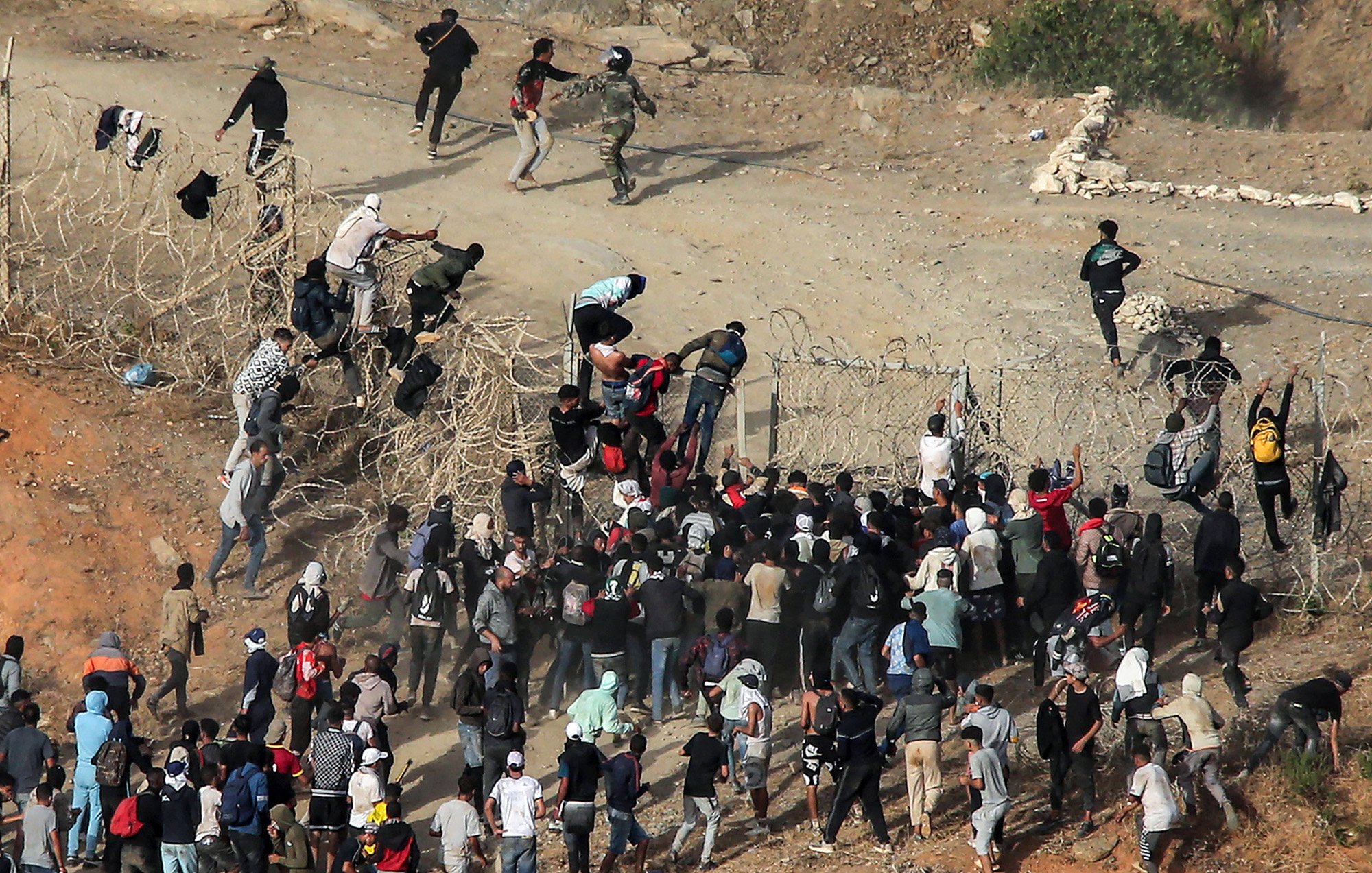 L'image montre un groupe de personnes tentant de franchir une barrière ou une clôture. On peut voir des individus escalader la clôture, tandis que d'autres semblent se rassembler en dessous. Il y a également des agents de sécurité présents à proximité, semblant surveiller la situation. L'environnement est celui d'une zone de passage, avec des éléments de terrain et des groupes de personnes manifestement en mouvement. L'atmosphère semble tendue, suggérant une situation d'urgence ou de conflit.