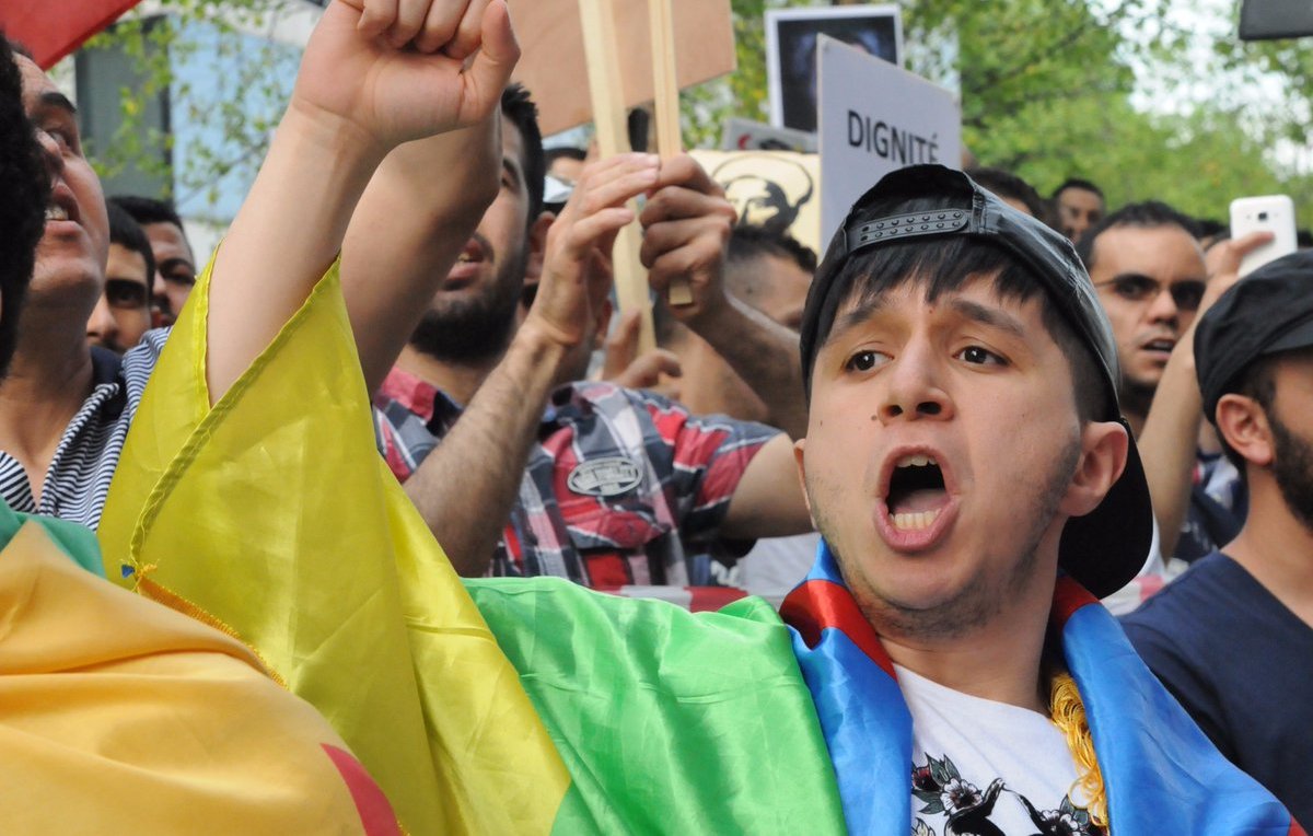 L'image montre une scène de manifestation, où un jeune homme, portant un drapeau coloré, crie avec passion. Il semble mobilisé et engagé dans la cause pour laquelle les manifestants se sont rassemblés. En arrière-plan, on peut voir d'autres personnes tenant des pancartes et exprimant leurs préoccupations. L'atmosphère est remplie d'énergie et de détermination.