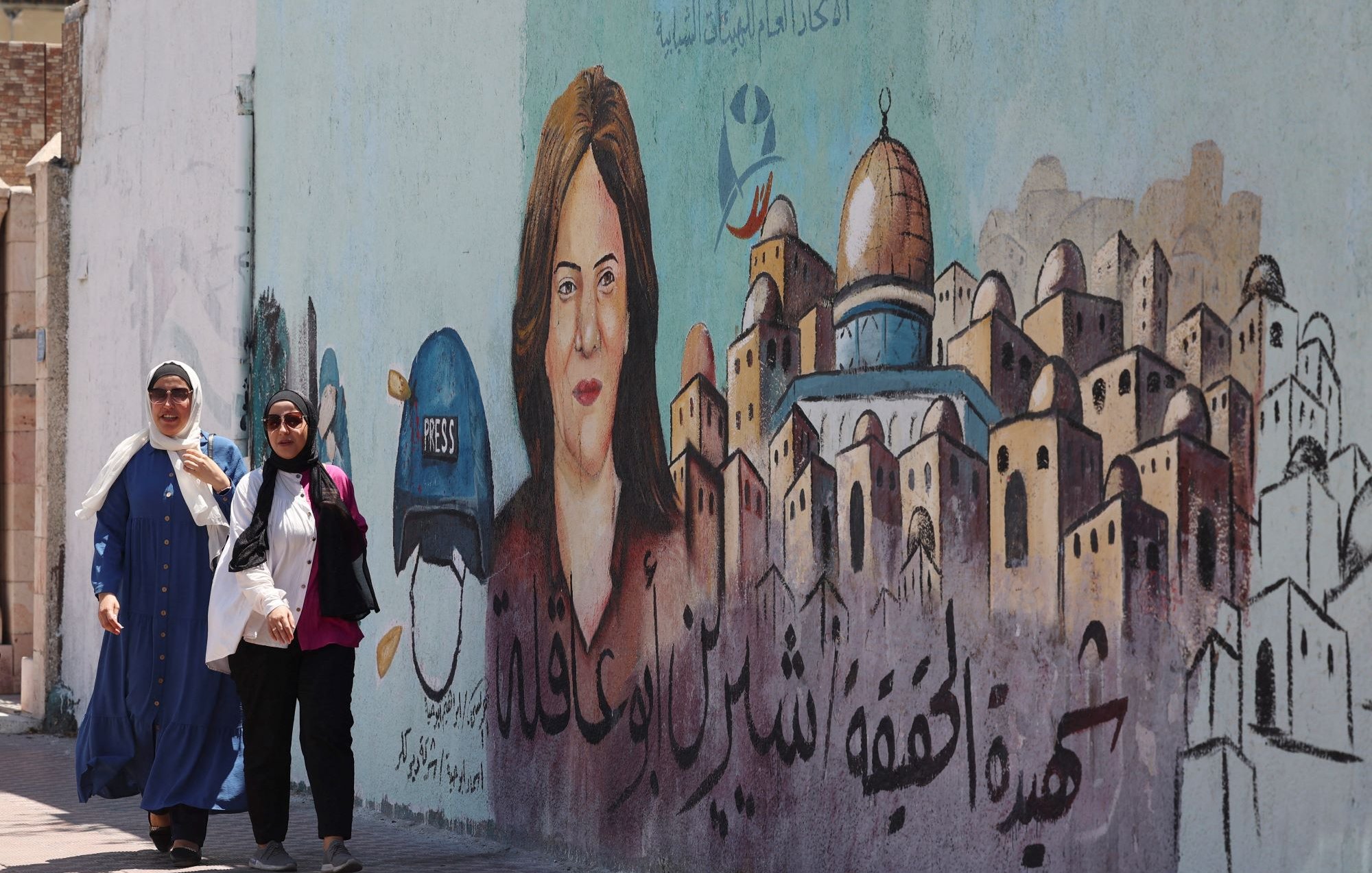 L'image montre deux femmes marchant le long d'un mur décoré d'une fresque. La fresque représente le portrait d'une femme avec un casque portant l'inscription "PRESS", suggérant qu'elle est journaliste. En arrière-plan, on aperçoit des maisons stylisées qui pourraient évoquer une ville ou un quartier particulier. Le texte en arabe est également visible, mais seul le contexte artistique et le message de solidarité ou de mémoire peuvent être interprétés. L'atmosphère est animée et engagée.