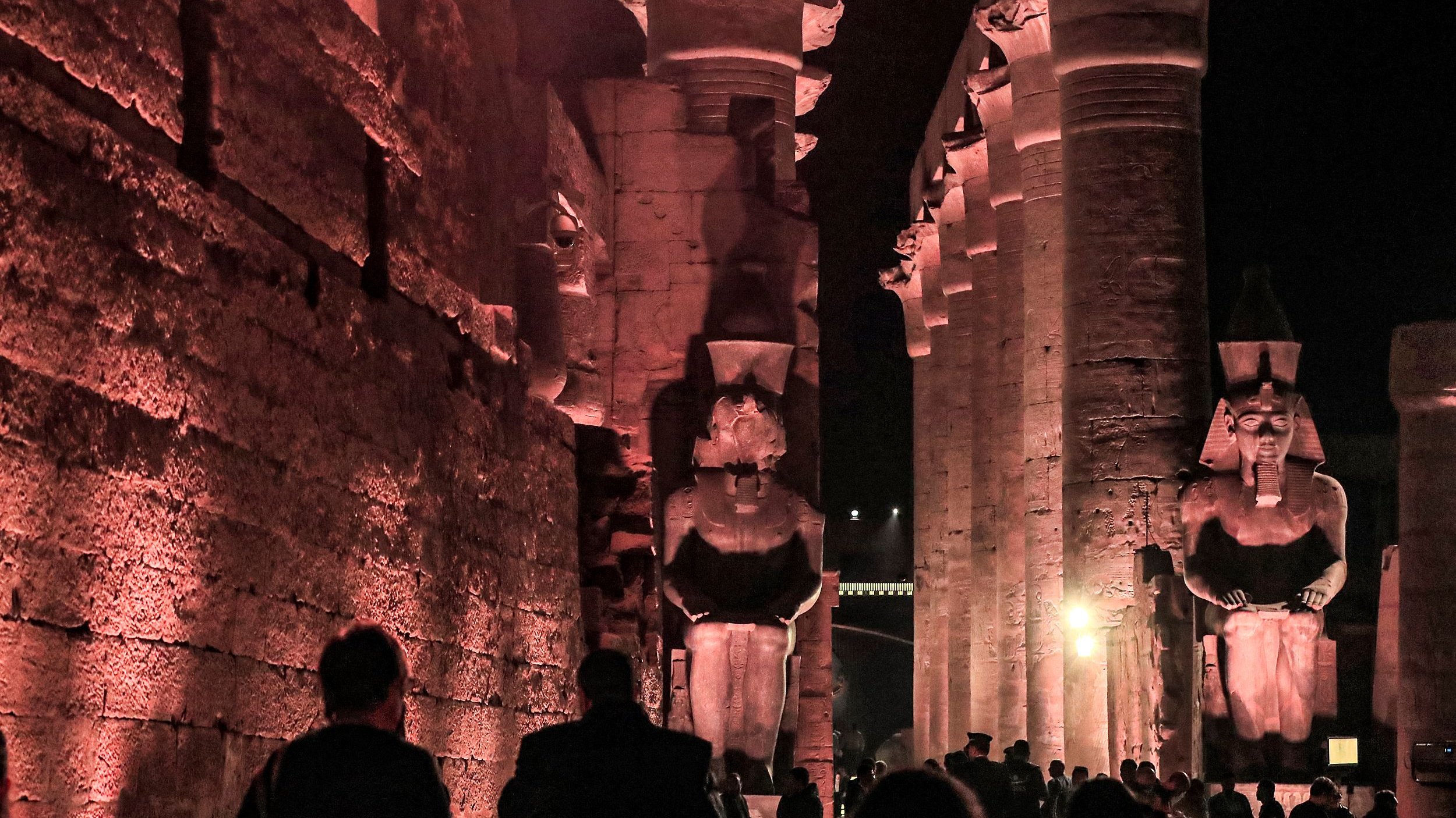 L'image présente un site historique, probablement des ruines égyptiennes, éclairées par des lumières tamisées, ce qui crée une atmosphère mystérieuse. On peut voir de grandes statues de pharaons ou de divinités, flanquant une structure impressionnante faite de colonnes. Les visiteurs se déplacent à travers le site, ajoutant une dimension humaine à cette scène ancienne, où l'architecture et les sculptures témoignent de la grandeur de la civilisation égyptienne. Les ombres et la lumière jouent un rôle important, accentuant les détails des sculptures et des murs.
