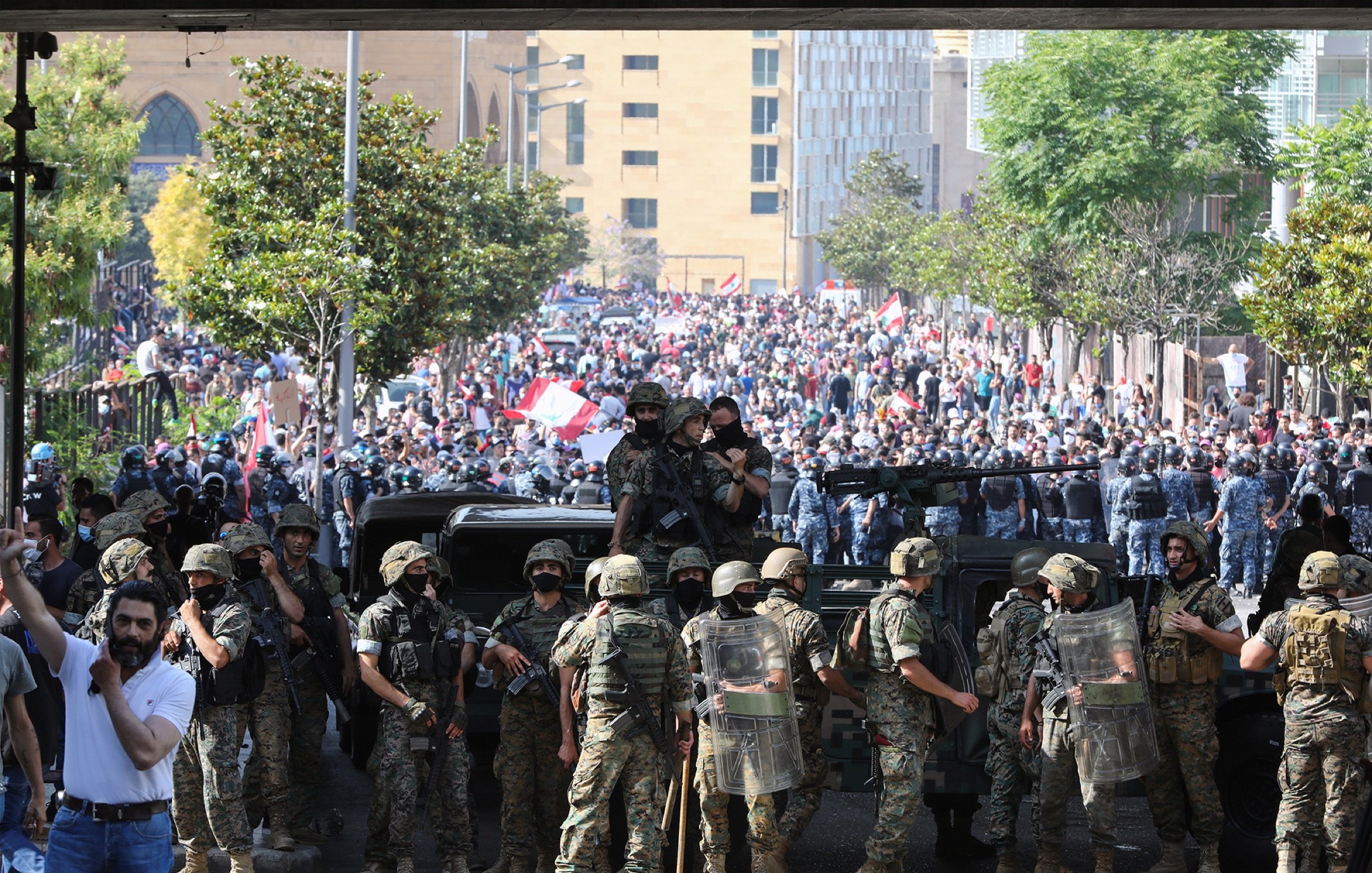 L'image montre une scène de protestation dans une ville, probablement à Beyrouth. Au premier plan, des soldats en uniforme sont alignés, portant des équipements de protection et des casques. Ils semblent faire face à une grande foule de manifestants qui se trouvent derrière eux. La foule est chargée d'énergie et porte des drapeaux, ce qui indique qu'il s'agit d'une manifestation pour une cause. Au fond, on peut voir des bâtiments urbains, ce qui ajoute un contexte à la scène. L'atmosphère est tendue, reflétant un moment significatif dans une dynamique sociale ou politique.