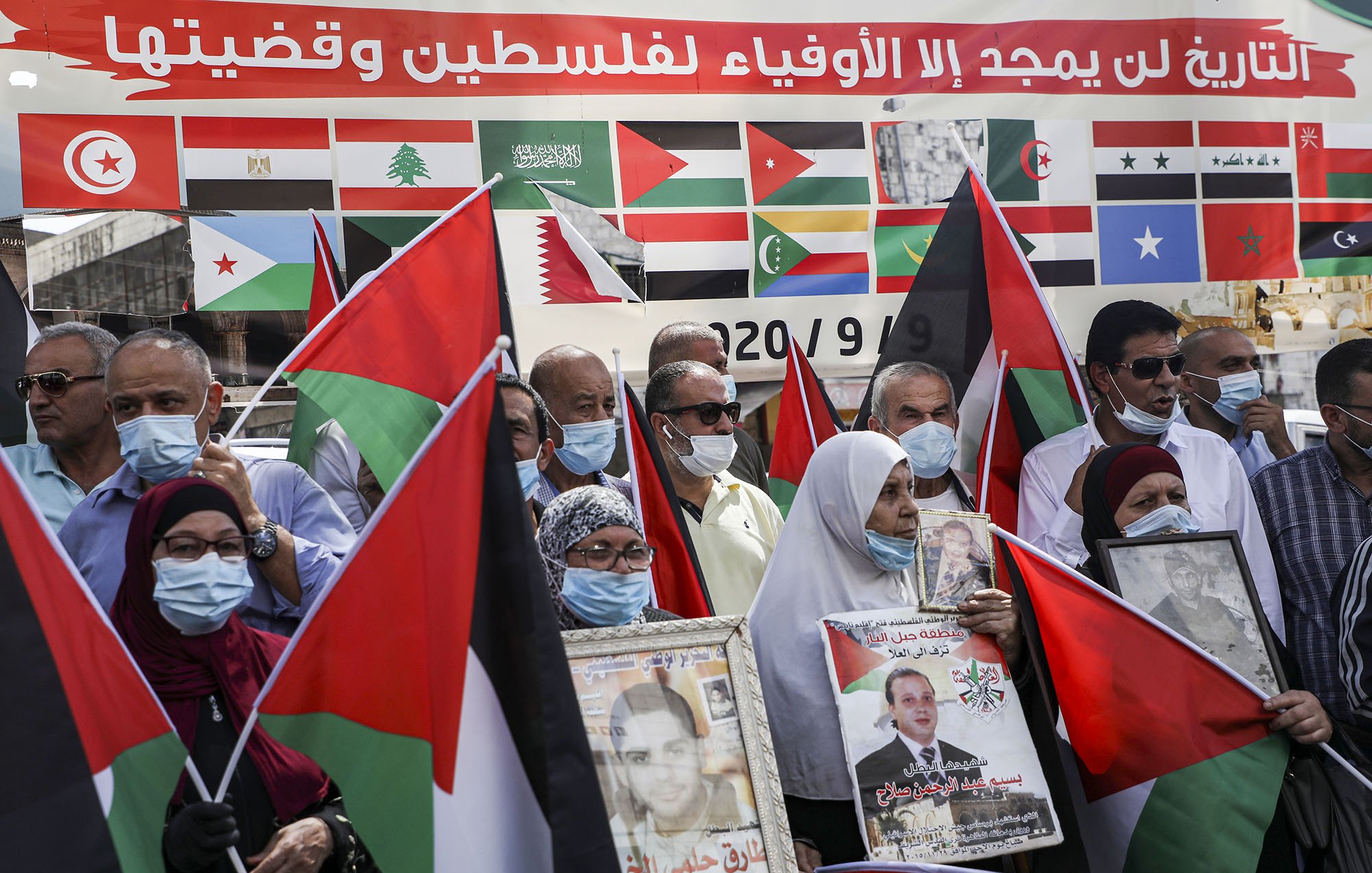 L'image montre un groupe de personnes rassemblées, portant des masques et tenant des drapeaux. Ils semblent manifester pour soutenir la Palestine, avec des affiches et des portraits en mains. En arrière-plan, on peut voir une grande bannière ornée de multiples drapeaux de différents pays. L'ambiance est engagée et solidaire, témoignant d'une mobilisation pour une cause politique.