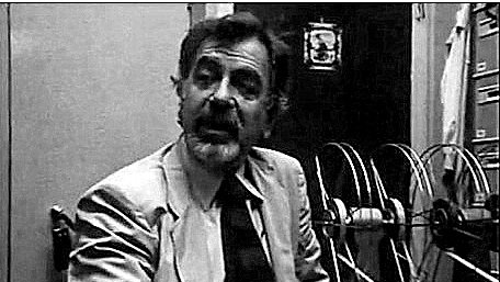 L'image présente un homme assis dans un bureau. Il porte un costume clair et une cravate foncée. Son expression faciale semble animée, presque passionnée, comme s'il était en train de discuter ou d'expliquer quelque chose. À l'arrière-plan, on peut voir des bobines de film ou des documents, ce qui donne une impression d'environnement de travail lié à la production ou à la recherche. L'éclairage est sombre, créant une ambiance vintage, typique des années passées.