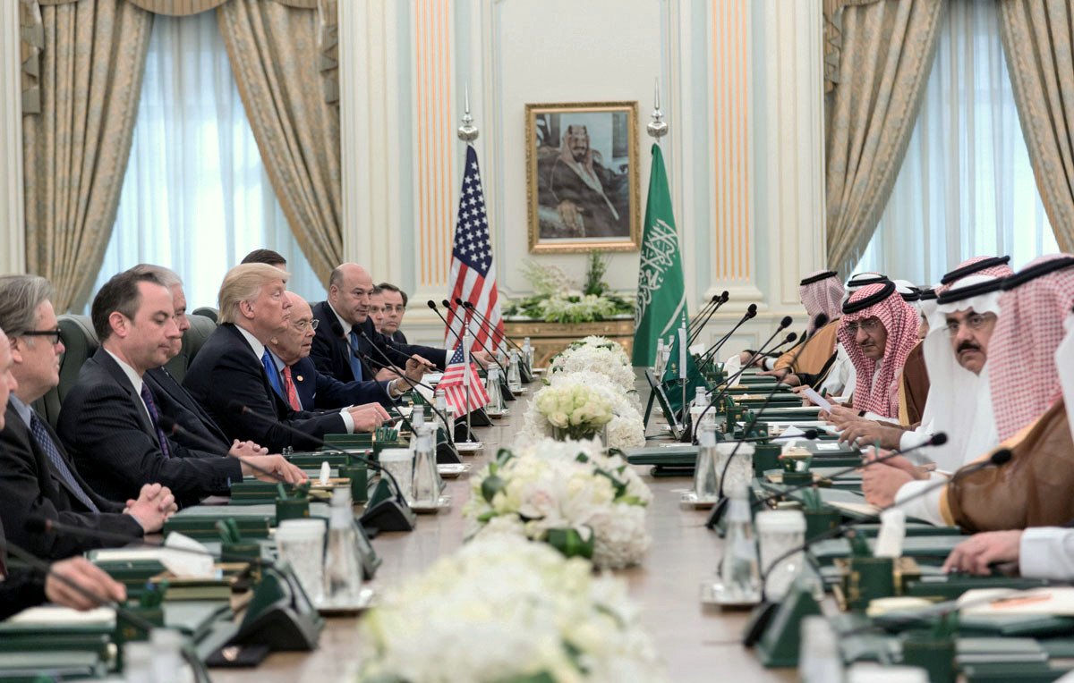 L'image montre une réunion formelle dans une grande salle avec une longue table. D'un côté, plusieurs représentants américains, dont on peut distinguer deux hommes en costume, ainsi qu'une personne qui semble être le président. En face d'eux, des dignitaires saoudiens, également en costume traditionnel. La salle est ornée de drapeaux américain et saoudien, et des arrangements floraux décoraient la table. L'atmosphère semble sérieuse et collaborative.