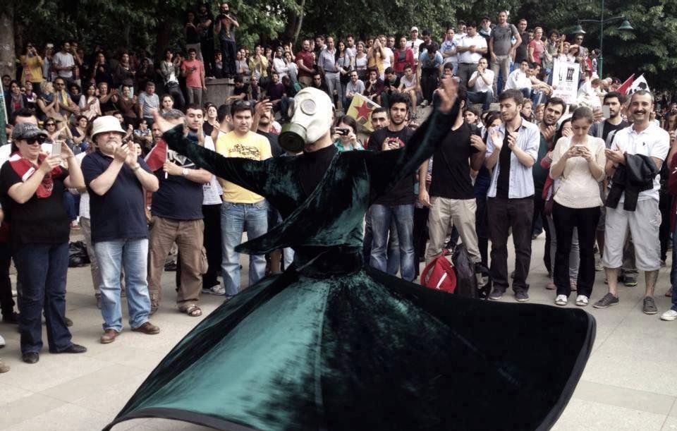 L'image montre une scène de rassemblement en plein air où une personne vêtue d'une longue robe vert foncé tourne gracieusement. Cette personne porte un masque à gaz, ce qui ajoute une dimension étrange et intrigante à la performance. Autour, une foule observe, certains prenant des photos avec leurs téléphones. L'ambiance semble à la fois festive et solennelle, avec des expressions variées parmi les spectateurs. Les arbres en arrière-plan ajoutent une touche de nature à la scène.
