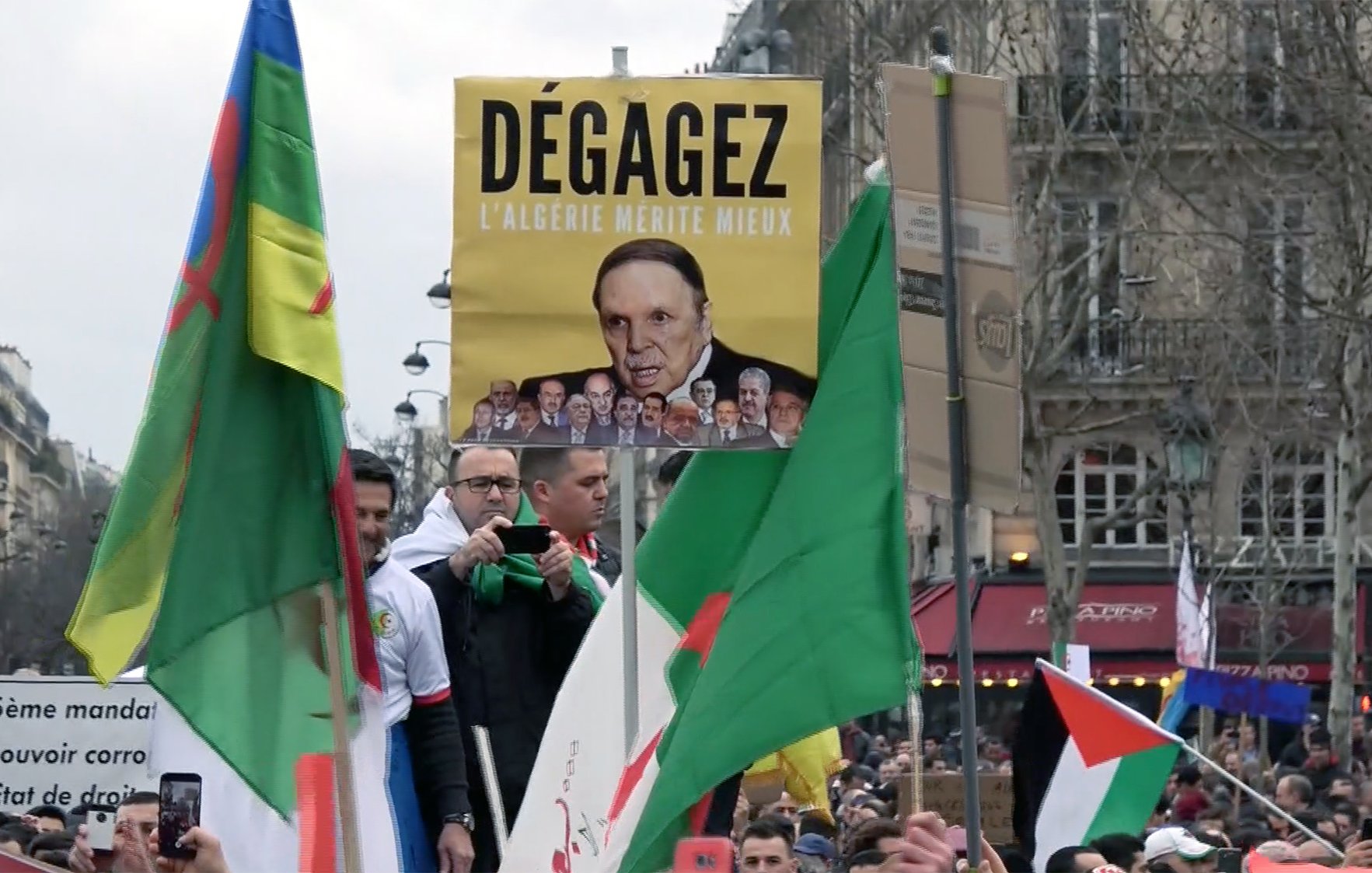 L'image montre une manifestation avec des participants portant des drapeaux. Au centre, un homme tient une grande pancarte sur laquelle est écrit "Dégagez l'Algérie mérite mieux", accompagnée d'une photo d'une personne. Les manifestants semblent exprimer des opinions politiques, et l'atmosphère semble être celle d'une contestation pacifique. Des slogans sur les pancartes reflètent des revendications pour des changements en Algérie.