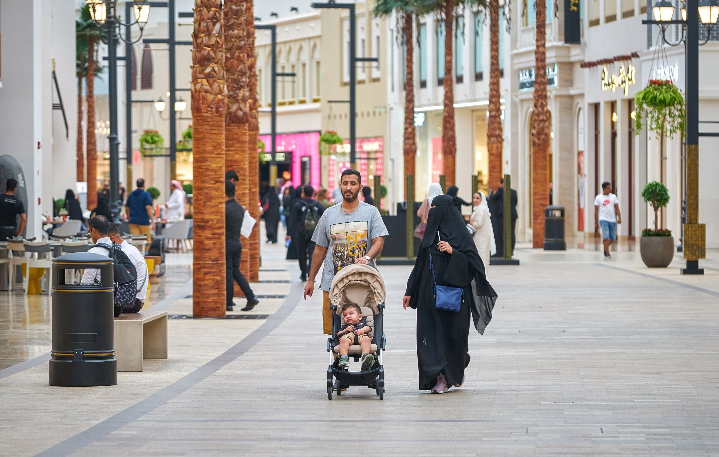 L'image montre une scène animée dans un centre commercial. On peut voir des palmés en arrière-plan et des magasins de différentes boutiques. Un homme marche avec un enfant dans une poussette, tandis qu'une femme, vêtue d'une abaya, les accompagne. Plusieurs autres personnes sont visibles dans l'allée, créant une ambiance de vie urbaine dynamique. La lumière est claire et l'atmosphère semble accueillante.