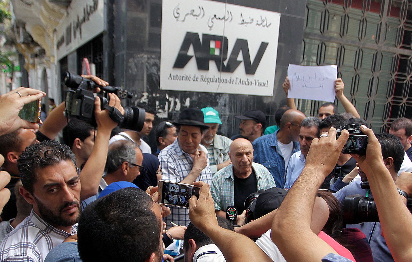 L'image montre un groupe de personnes rassemblées autour d'un homme au premier plan, visiblement en pleine discussion ou confrontation. Plusieurs caméras et téléphones portables sont levés, capturant l'événement. En arrière-plan, on peut voir une bannière avec le mot "ARAV" qui fait référence à l'Autorité de Régulation de l'Audiovisuel. La scène semble être un moment de protestation ou de manifestation, avec des expressions variées sur les visages des participants.