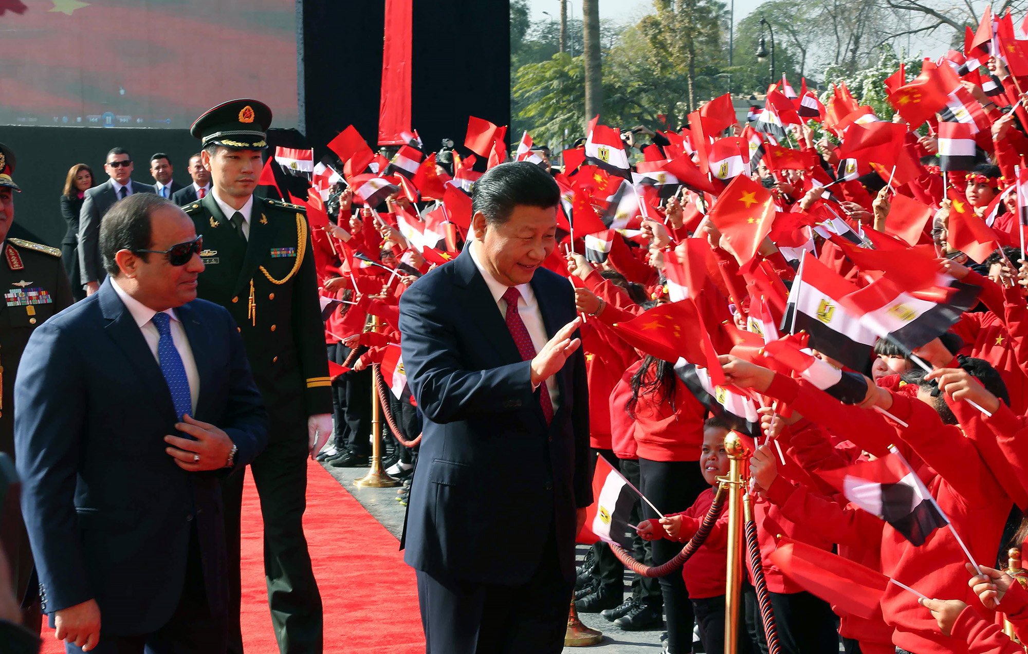 L'image montre un événement public avec des drapeaux de la Chine et de l'Égypte. Au premier plan, deux figures politiques semblent interagir avec la foule. L'un des hommes porte un costume sombre et des lunettes de soleil, tandis que l'autre est habillé d'un costume clair et sourit en saluant les personnes présentes. La foule, vêtue de vêtements rouges, tient des drapeaux et semble enthousiaste. On peut voir un tapis rouge et des gardes en uniforme à l'arrière-plan. L'ambiance est festive et accueillante.