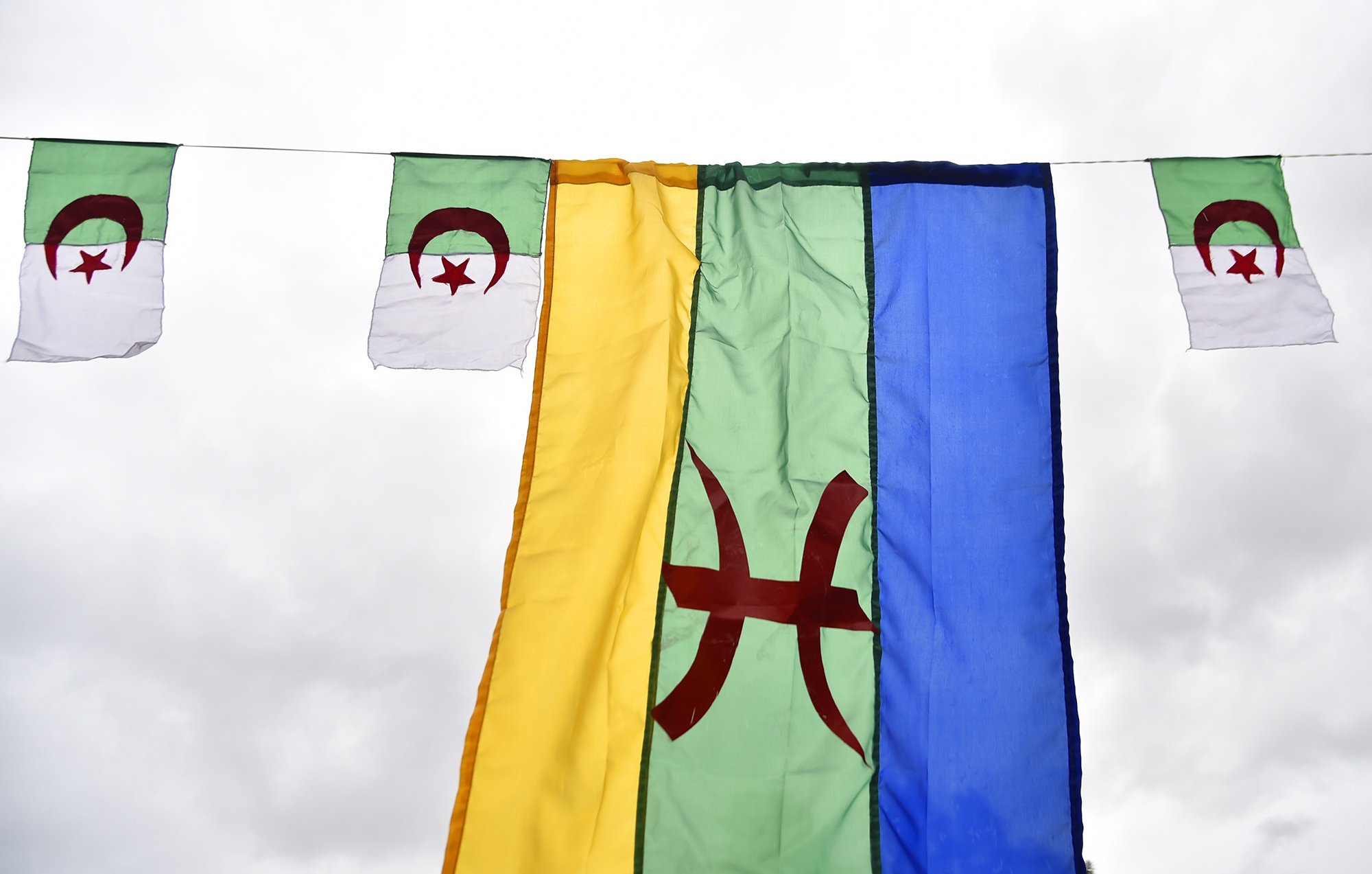 L'image montre plusieurs drapeaux suspendus. Au centre, il y a un grand drapeau de couleur verte avec un symbole rouge au centre, probablement représentant le mouvement amazigh. De chaque côté, il y a de plus petits drapeaux, qui semblent être le drapeau algérien, avec les couleurs verte et blanche et le croissant et l'étoile rouges. L'ensemble est accroché sous un ciel nuageux, créant une ambiance festive ou symbolique.