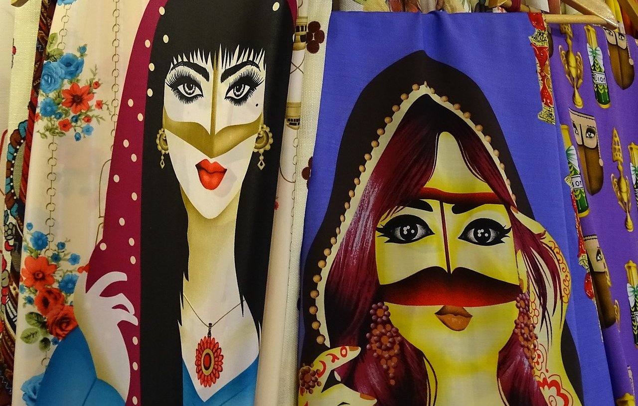 L'image présente deux illustrations de femmes, affichées sur des tissus. À gauche, une femme porte un foulard avec des dessins floraux, des yeux accentués et une bouche rouge. À droite, une autre femme arbore des bijoux traditionnels et un voile coloré, avec un visage stylisé et des traits distinctifs. Les deux illustrations ont un style graphique vibrant et coloré, avec des motifs qui ajoutent à leur allure artistique.